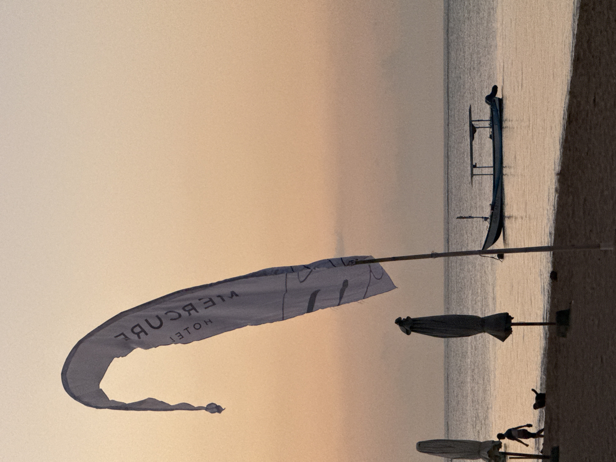 A medium shot captures a beach scene at dusk, likely in Denpasar, Indonesia. In the foreground, a tall, white flag with dark lettering, possibly indicating a hotel or resort, billows on a pole against a gradient sky of soft oranges and yellows. The flag is partially obscured, with its top curved and its lower part trailing. To the left, two closed beach umbrellas stand on the sand, their dark fabric contrasting with the lighter beach. A silhouetted figure of a person walks along the shoreline, accompanied by a silhouetted dog. In the middle ground, a small boat with a canopy floats on calm, reflective water. The sea stretches to the horizon, where it meets the fading light of the sky. The beach itself appears sandy and gently slopes towards the water.