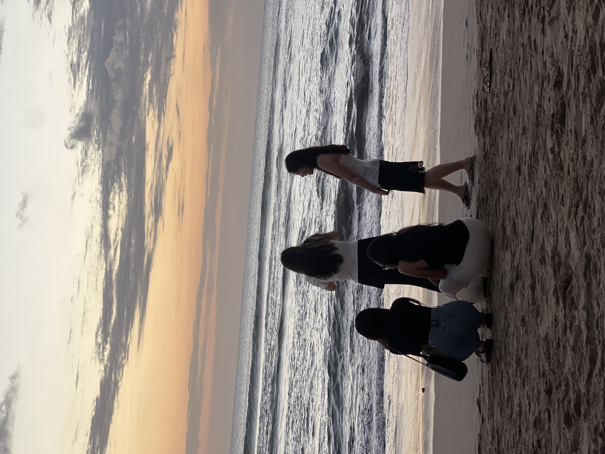 Four women are silhouetted against the horizon on a sandy beach in Kuta, Indonesia, during what appears to be sunset. The sky is a gradient of soft orange and yellow, with streaks of gray clouds. The ocean is calm, with small waves rolling onto the shore.
The woman on the far left is crouched down, facing the ocean, with a black purse at her side. She is wearing a black top and blue jeans.
Next to her, a second woman is also crouched, facing away from the viewer and towards the ocean. She is wearing a black top and light-colored pants.
A third woman stands facing the ocean, with her back to the camera. She is wearing a white short-sleeved top and a black skirt.
On the far right, a fourth woman stands, facing towards the left of the frame and looking out at the ocean. She is wearing a white strapless top and a black skirt. Her hair is long and dark.
The sand in the foreground is coarse and textured, with a few scattered seashells. The overall atmosphere of the image is serene and peaceful.
