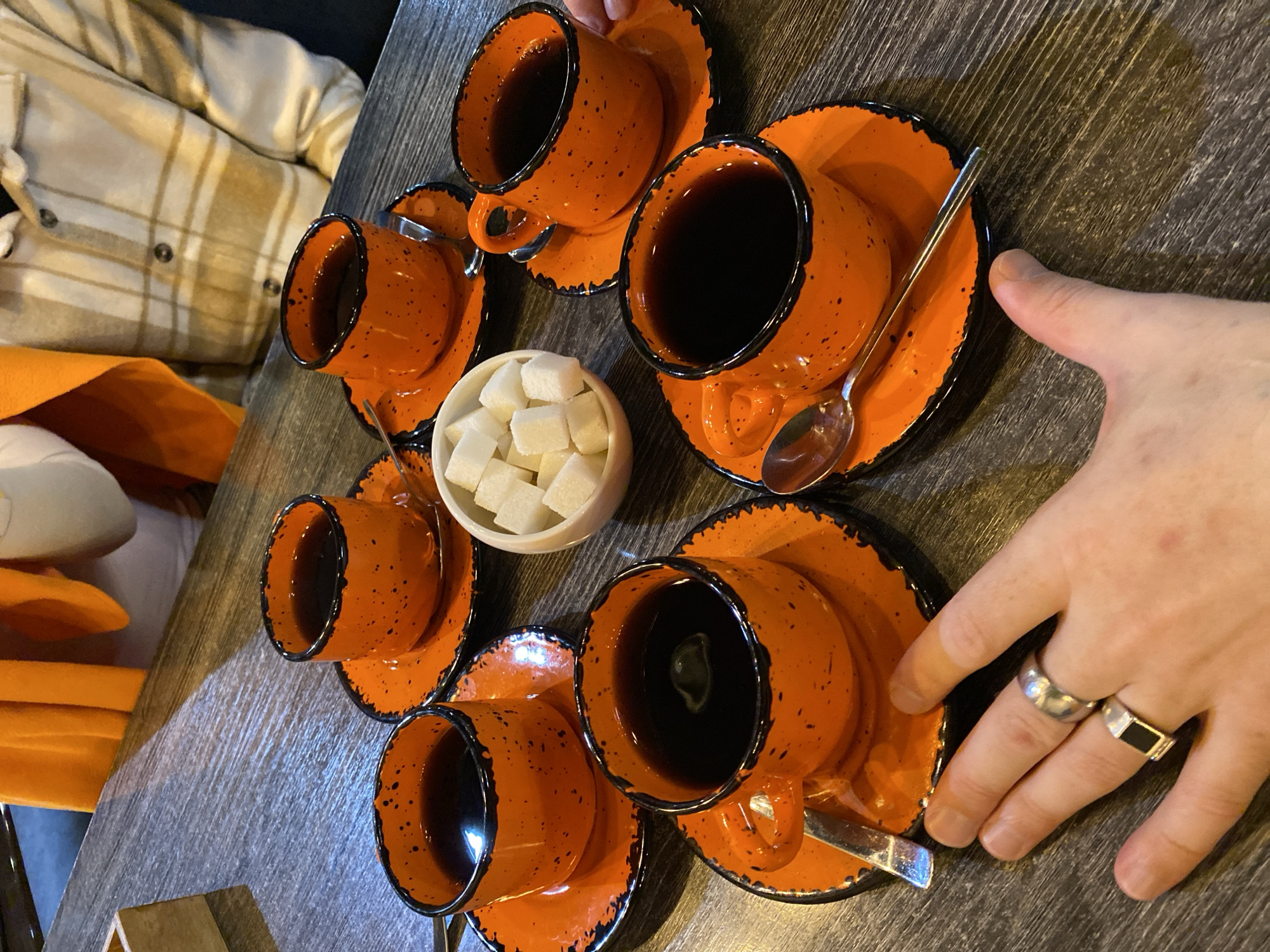 A close-up, high-angle shot displays a collection of six orange coffee cups with black rims and speckles, each sitting on a matching saucer, arranged in a semi-circular pattern around a small white bowl filled with sugar cubes. Each saucer holds a silver spoon. The cups are filled with a dark liquid, presumably coffee. The scene is set on a dark, possibly wooden, table. A person's hand wearing two rings (one silver and one with a black stone) is visible on the right side of the image, near one of the cups. Partial views of a person wearing a beige and white plaid shirt and orange garment appear in the upper left quadrant of the image. The location of the photograph is Izhevsk, Russia.