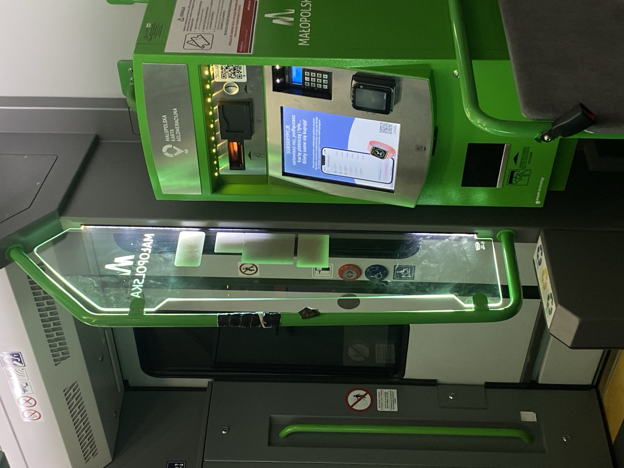 The image depicts the interior of a vehicle, likely a tram or bus, with a ticket vending machine on the right and a door on the left. The ticket machine is bright green and features a touchscreen display showing a smartphone and a smartwatch, with Polish text relating to subscriptions and purchasing tickets. Above the touchscreen, there are slots for tickets and payment, a QR code, and a keypad. The text "MAŁOPOLSKA KARTA AGLOMERACYJNA" is visible on the machine. To the left of the machine, a transparent vertical panel with the inverted text "MAŁOPOLSKA" is illuminated by green LEDs. On the far left, a gray door is visible with green handrails. The floor appears to be yellow in the foreground. The scene is well-lit, with the green lighting from the ticket machine and the vertical panel being prominent. The context suggests a public transportation setting in Kępa Bogumiłowicka, Poland, where this automated ticket vending system is operational. There are no people visible in the image.