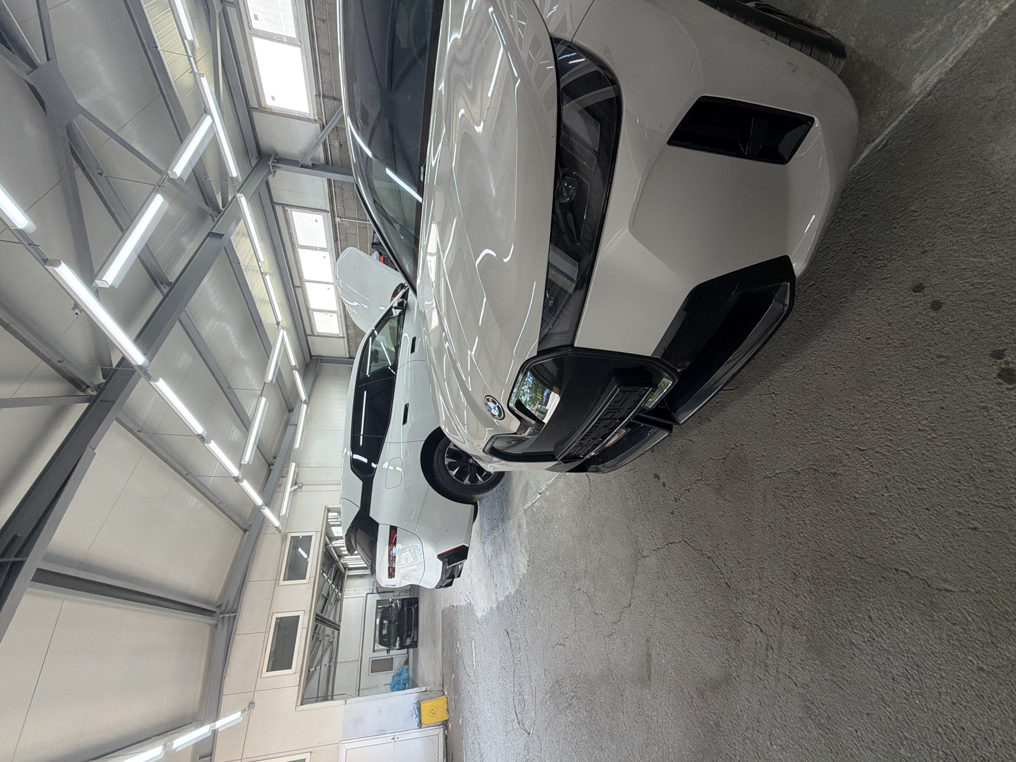 A two-car garage workshop is depicted in Burgas, Bulgaria. The scene is lit by fluorescent ceiling lights. Two white BMW iX electric SUVs are parked inside. The SUV in the foreground is positioned to the right, with its front right quarter facing the viewer, and its hood is closed. The second white BMW iX is parked behind the first, slightly to the left, with its rear right quarter visible and its hood is open. A black car is parked further in the background on the left. The floor is made of concrete, and the walls are constructed of brick and metal panels. Yellow trash bins and blue plastic bags are visible on the left side of the image. The overhead structure of the garage is made of metal beams.