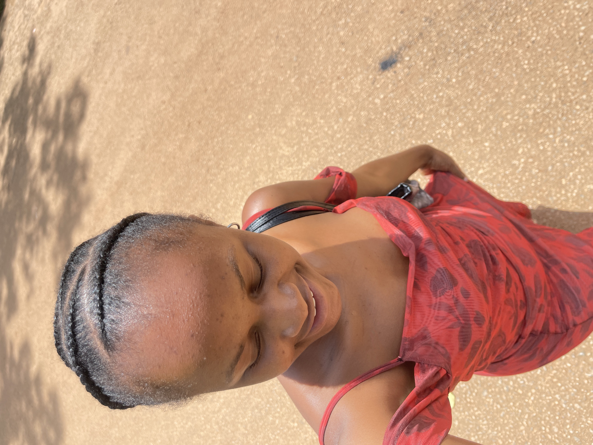 A young Black woman with her eyes closed and a slight smile is captured from a high-angle perspective, looking down. Her hair is styled in four neat braids. She wears an off-the-shoulder red mesh dress with a subtle floral pattern. A black purse with a silver buckle is slung over her right shoulder. The background is a sandy, light brown surface, possibly a path or unpaved road, with scattered shadows from trees overhead. The scene appears to be outdoors in daylight. The location is the city of Rafin Par, Nigeria.
