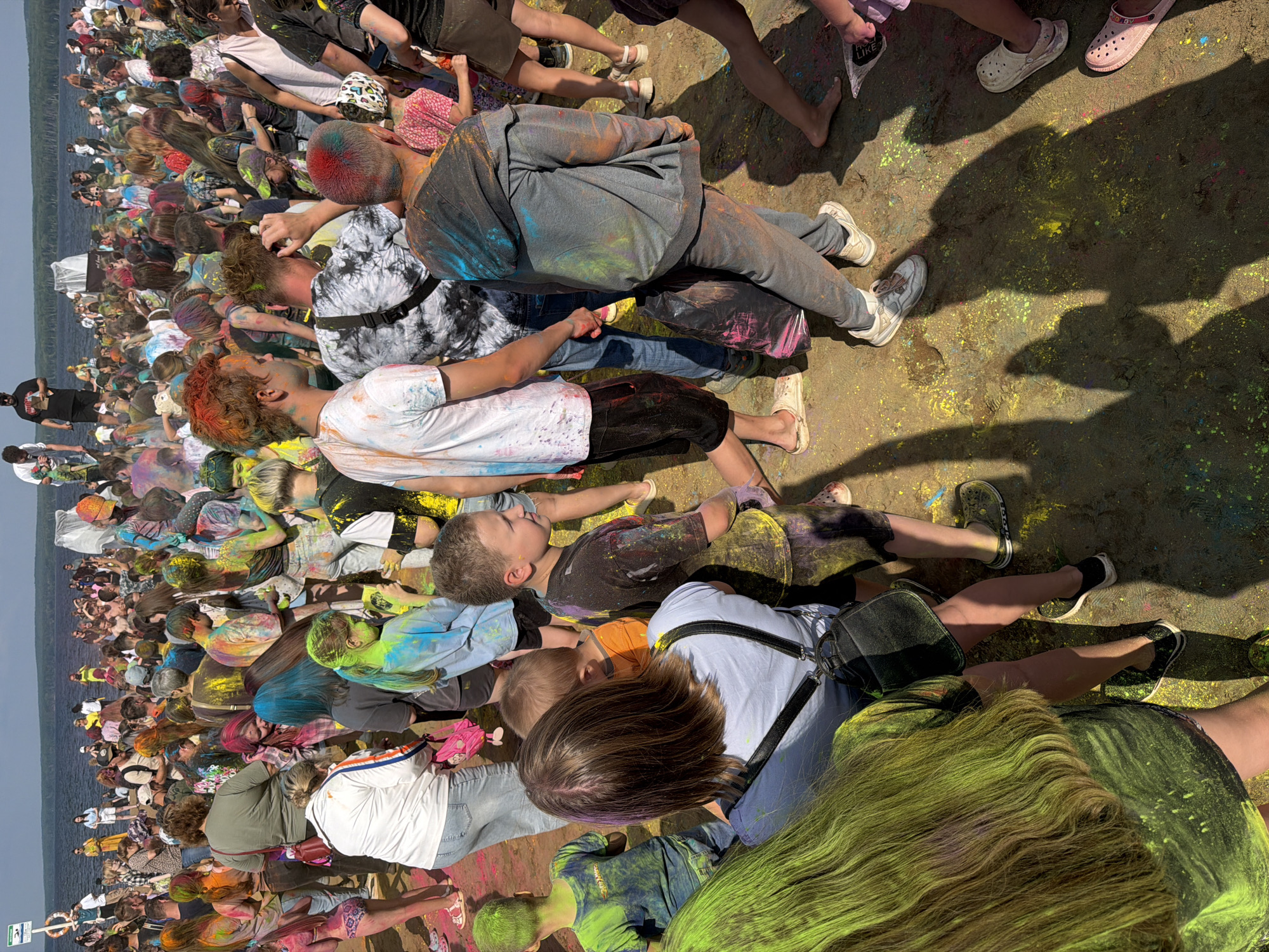 The image depicts a vibrant outdoor festival, likely a Holi celebration, taking place on a sandy area near a body of water. The ground is scattered with brightly colored powder, and many individuals are coated in the same festive hues. The crowd appears diverse in age, with children and adults mingling. People are dressed in casual clothing, with some wearing t-shirts, shorts, and jeans. The atmosphere seems lively and joyous, with participants actively engaging in throwing or having colored powder applied to them. In the background, the calm expanse of a lake or reservoir is visible, with a tree-lined shore in the distance. The sky is clear and bright, suggesting fair weather. This scene is consistent with the annual "Holi on the Kama" festival held in Chaikovsky, Russia, which traditionally takes place on the sandy beaches along the Kama River.