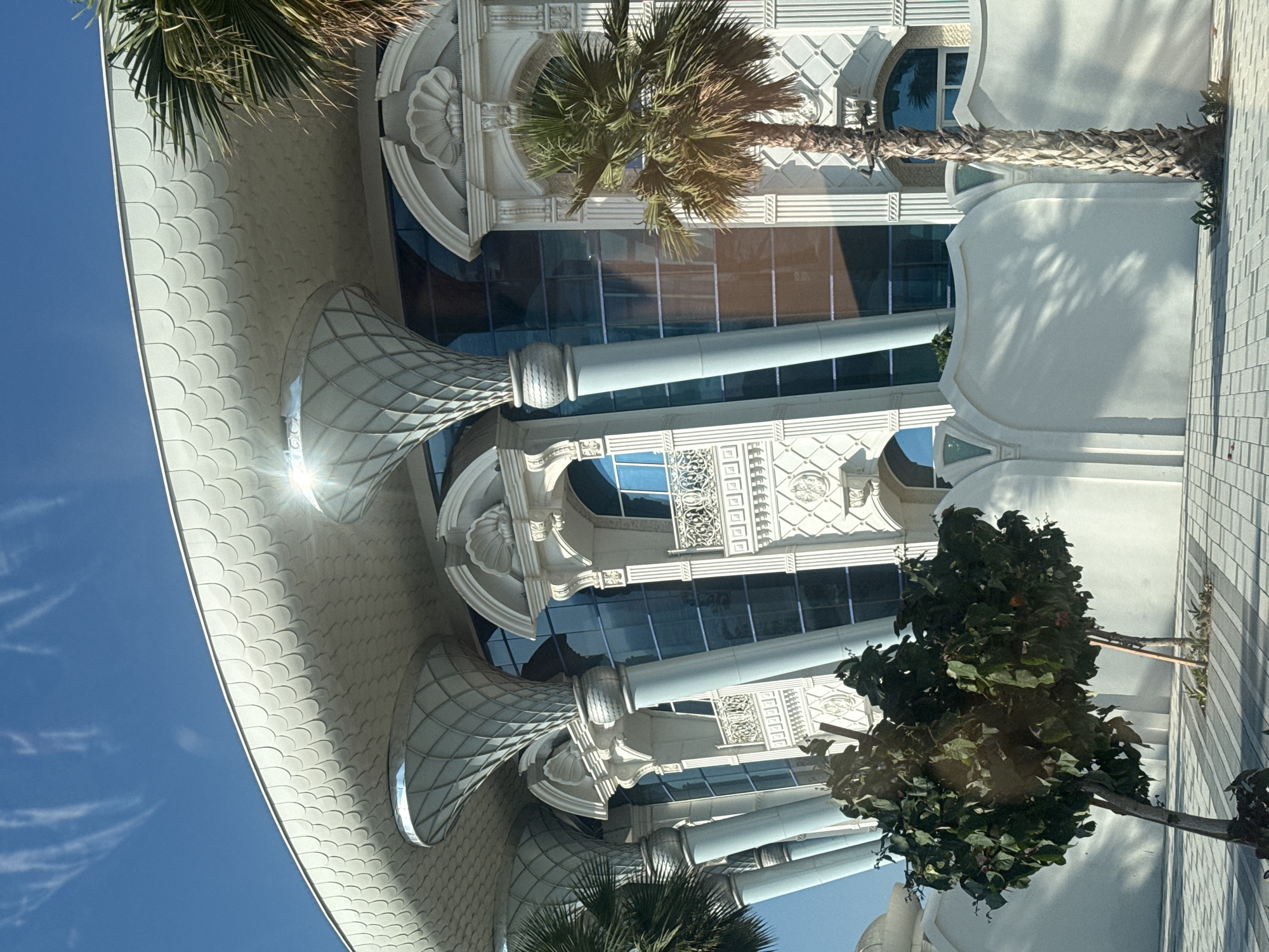 The image depicts a modern architectural structure with a white, scalloped roof supported by decorative, trumpet-shaped columns. The building features large, reflective blue glass windows and ornate white detailing around arched windows and balconies. Palm trees and a leafy tree are visible in the foreground, along with a white, curved wall. The scene is set on a bright, sunny day with a clear blue sky and scattered clouds. The architectural style suggests a high-end commercial or residential building, possibly a hotel or a luxury complex, located in Dubai, United Arab Emirates. The overall impression is one of opulence and contemporary design.