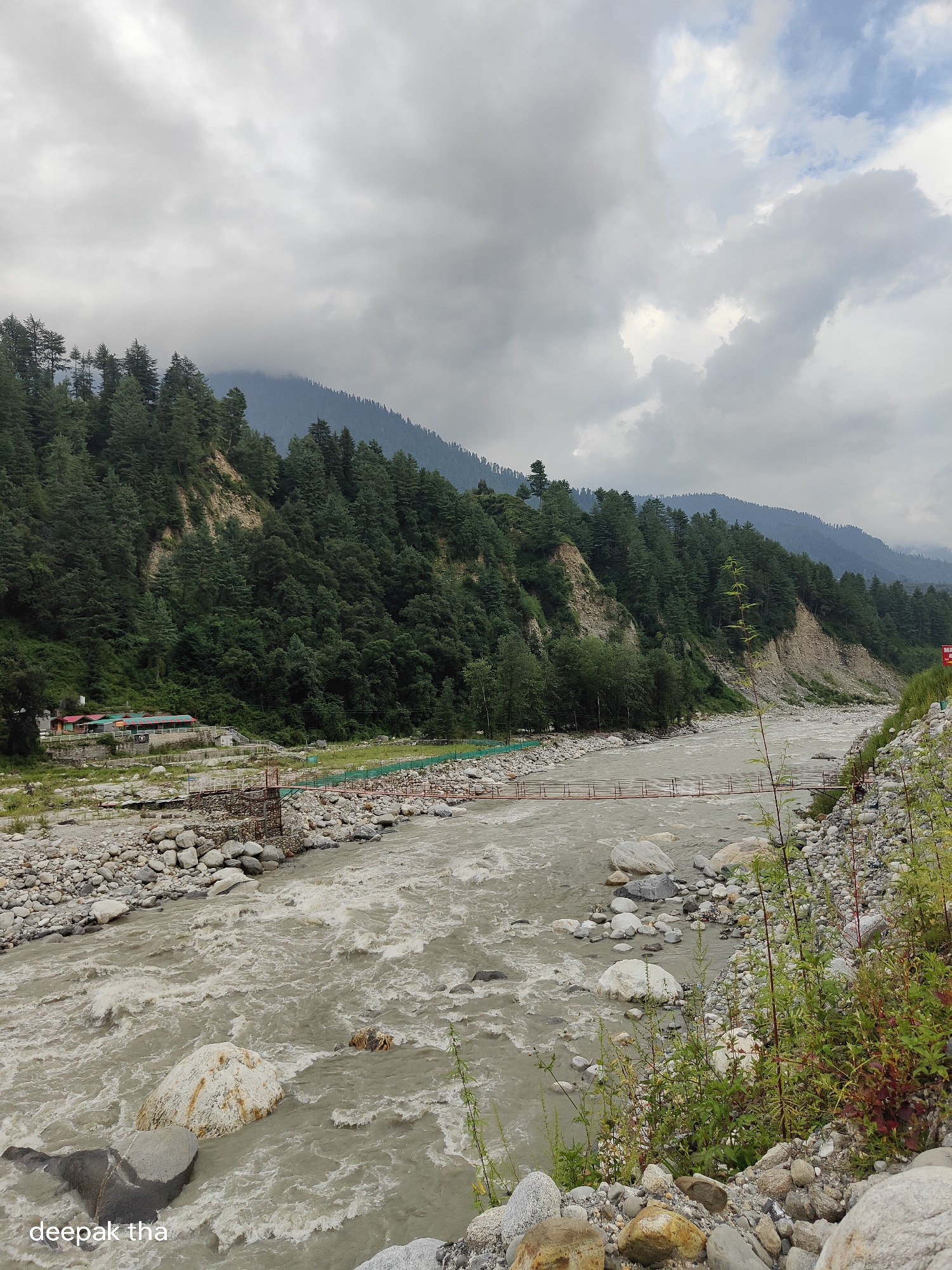 The image shows a wide, fast-flowing river with muddy water in Kalath, India. The riverbed is covered in large, smooth stones. On the left bank of the river, a lush green forest covers a steep hillside that rises into misty mountains. A small cluster of buildings with red roofs is visible nestled at the base of the hill near the river. A suspension bridge with a red railing crosses the river in the mid-ground. The sky is overcast with heavy, gray clouds. The overall impression is a rugged, natural landscape under a cloudy sky.