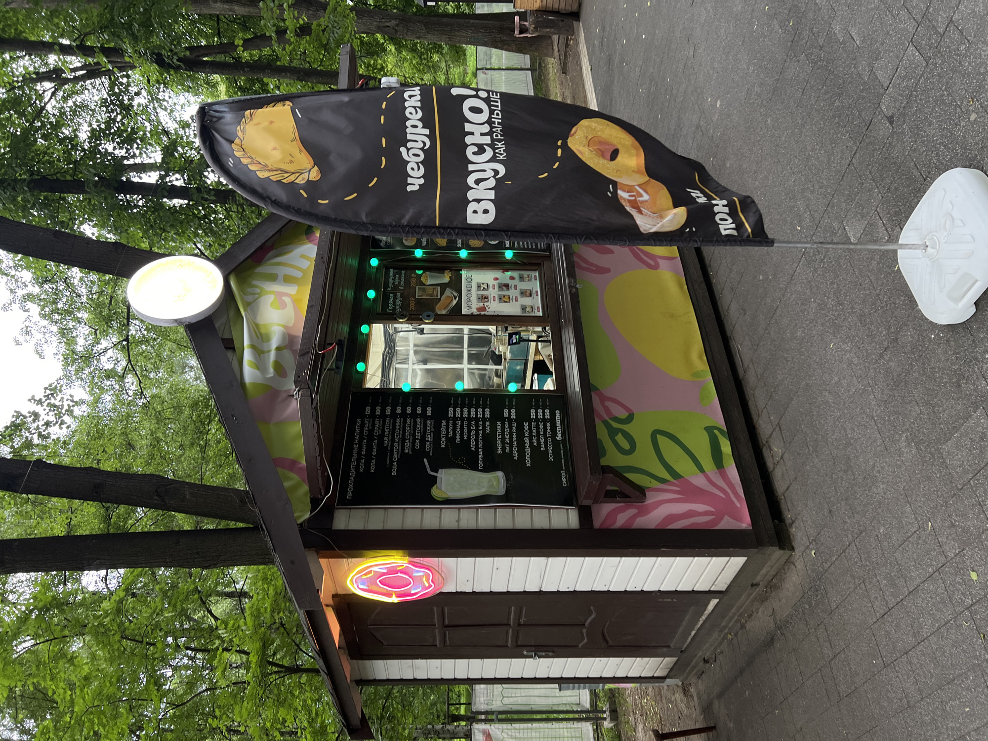 A street food kiosk stands in Mytishchi, Russia. The kiosk has a dark brown roof with a bright yellow light fixture attached. The walls are white paneling on the lower section and decorated with a pink and green leaf design on the upper section. A neon pink donut sign is mounted near the door. A black menu board is visible, listing a variety of drinks in Russian, including Coca-Cola, Fanta, Sprite, juice, lemonade, and coffee. A black teardrop-shaped advertising flag stands next to the kiosk, displaying a cartoon of a fried dough snack, likely a cheburek, and the words "ВКУСНО! КАК РАНЬШЕ" (Delicious! Like before). The flag is attached to a white plastic base on the ground. The kiosk is positioned on a paved area surrounded by green trees.