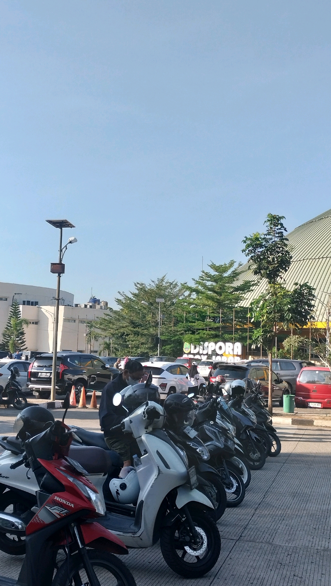 The image shows a parking lot in front of a building with a large "DISPORA" sign. The building appears to be a sports or recreational facility, possibly part of the Gelora Bandung Lautan Api (GBLA) complex, given the architectural style and the "DISPORA" signage which is often associated with sports and youth departments in Indonesia. The parking lot is filled with many motorcycles, parked in rows. In the foreground, several people are seen interacting with their motorcycles. One person, wearing a dark jacket and a face mask, is sitting on a white scooter. Another person is standing near a red motorcycle with "HONDA" branding. In the background, a row of cars are parked, and a solar-powered street light stands tall. Trees and a large, modern-looking building with a curved roof are visible behind the parking lot, suggesting an urban or semi-urban setting. The sky is clear and blue, indicating good weather.