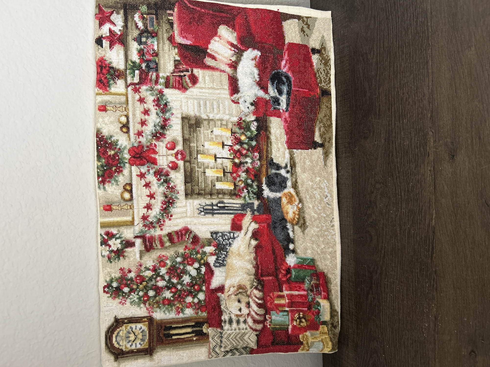 The image is a needlepoint tapestry depicting a cozy Christmas scene. In the center, a fireplace is adorned with garlands, stockings, and candles. To the left of the fireplace is a decorated Christmas tree with presents beneath it, and a grandfather clock. To the right of the fireplace is a red armchair with a small ottoman in front of it. Several animals are depicted in the scene: a golden retriever lies on a red sofa, a black and white cat sleeps on the ottoman, and a white dog stands on the red armchair. The tapestry is placed on a dark wooden surface. There is no indication of the specific location within Colorado Springs, United States of America.