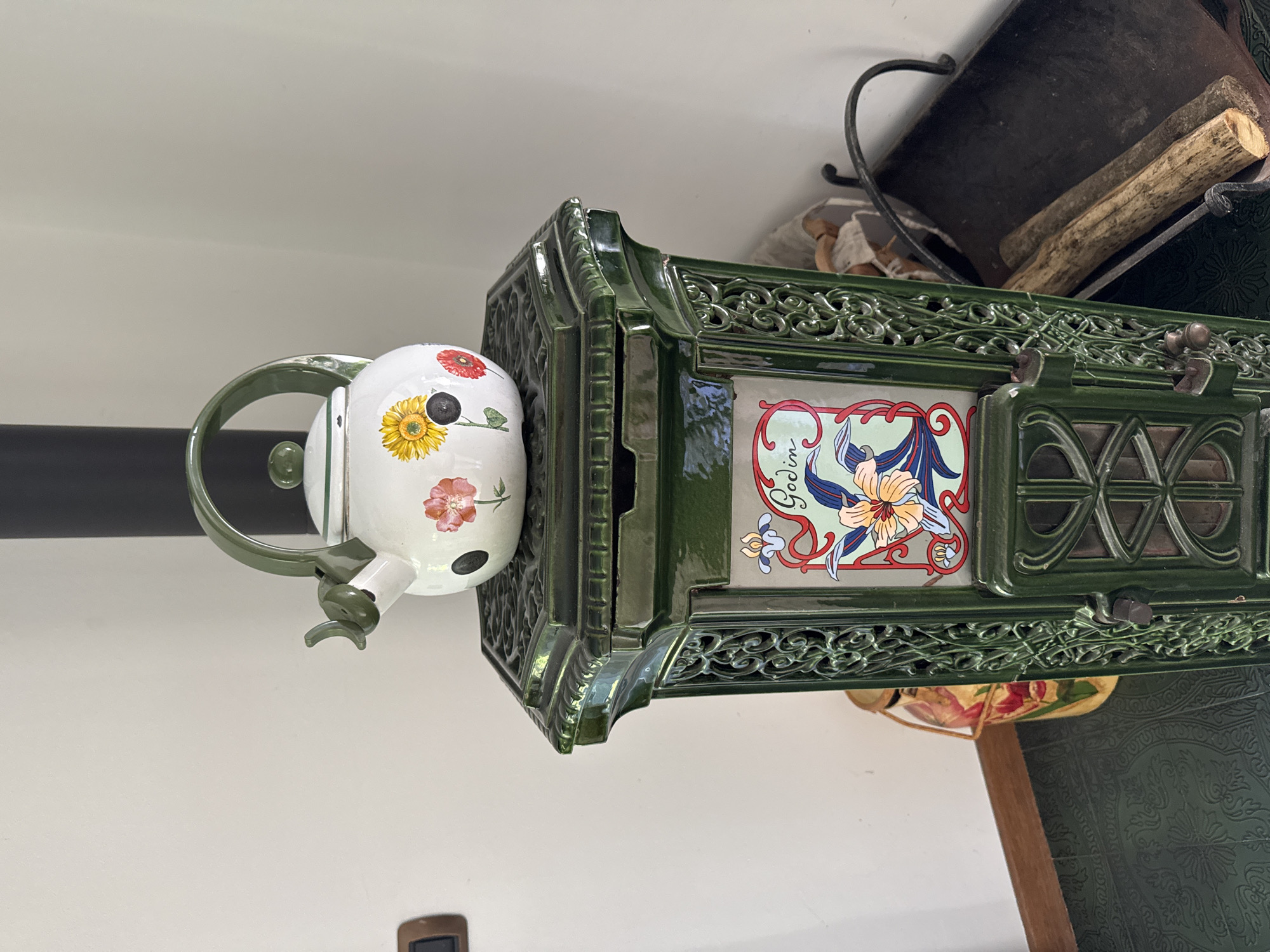 A close-up, eye-level shot captures a vintage green enamel stove with decorative ceramic tiles featuring a floral motif and the name "Godin." A white kettle with a green handle and floral designs sits atop the stove. To the left of the stove, a decorative ceramic vase with a floral pattern is partially visible. The stove is positioned on a patterned green floor. In the background, a black chimney pipe ascends vertically, and a metal ash pan with logs rests to the right of the stove. The wall behind the stove is painted a light color. The scene is likely an indoor setting, possibly a rustic or vintage-styled home. No people are present.