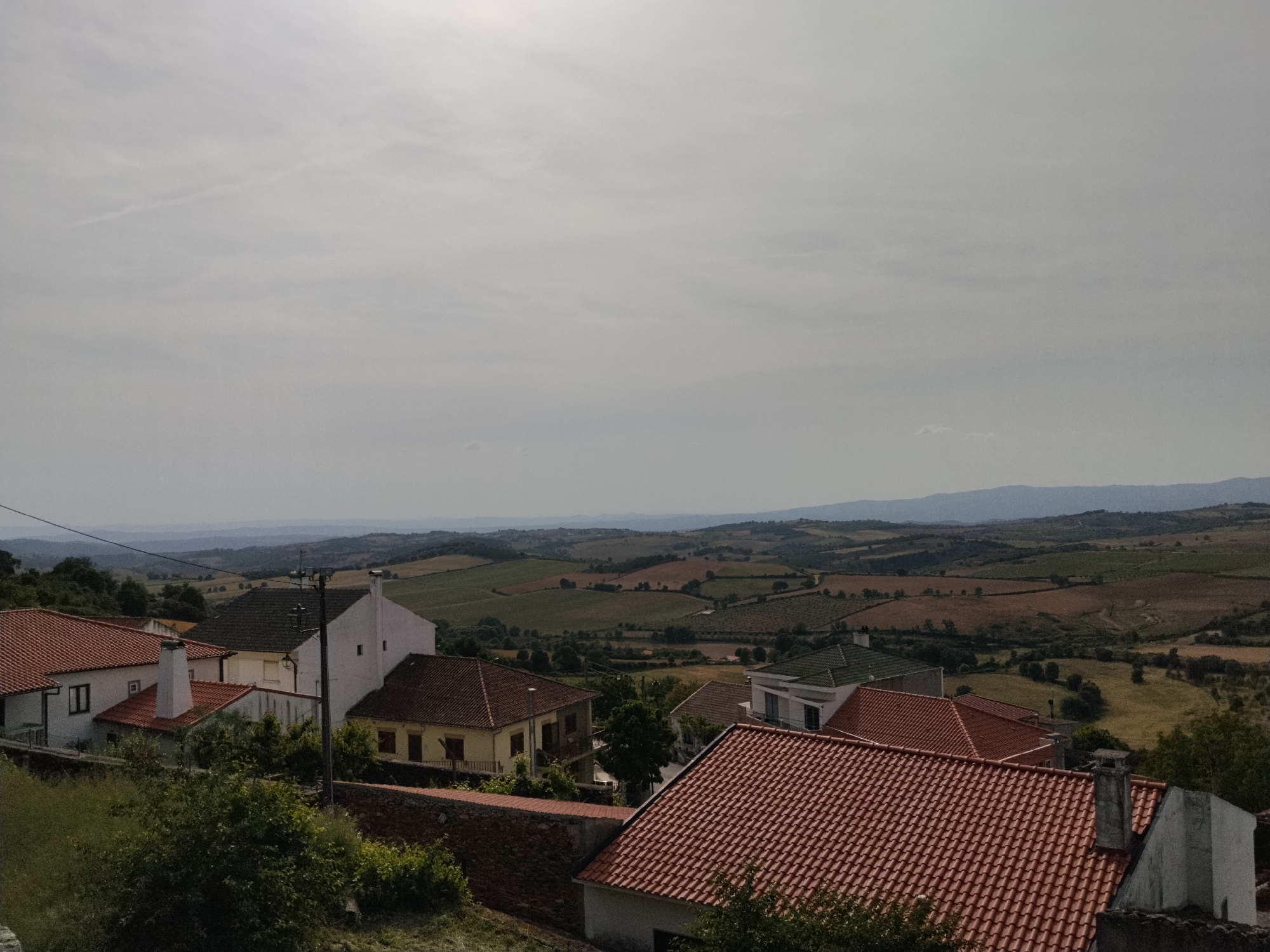 The image depicts a view of Mogadouro, Portugal, taken from an elevated perspective. In the foreground, tiled rooftops of several houses are visible, intermixed with green vegetation. A stone wall is present in the mid-ground. Beyond the buildings, rolling hills stretch into the distance, divided into agricultural fields of varying colors, suggesting different crops or stages of growth. A range of mountains can be seen on the horizon. The sky is overcast, with a uniform grey tone. An overhead powerline is visible in the upper left portion of the image.
