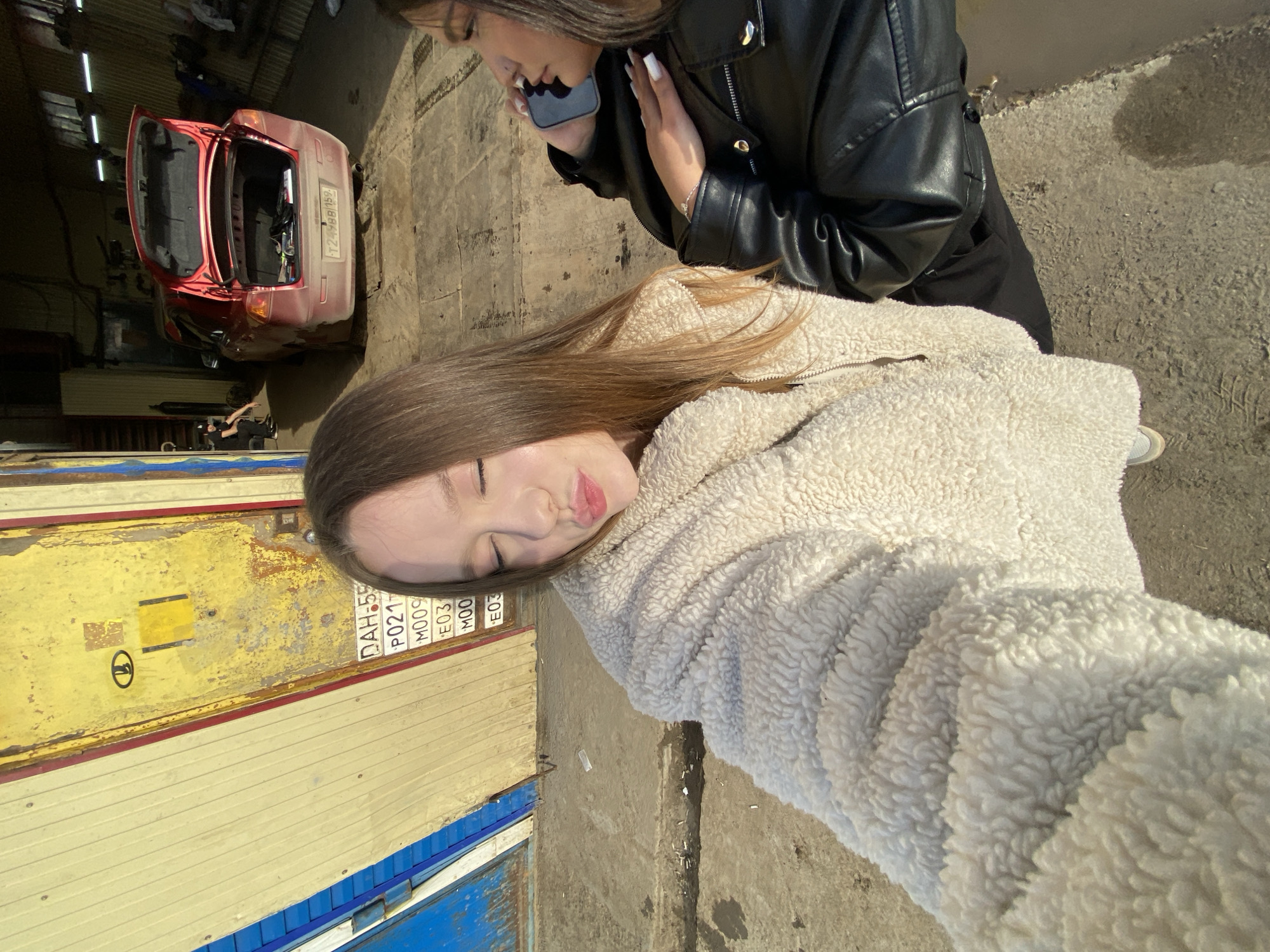 The image shows two women in what appears to be an auto repair shop in Chaikovsky, Russia. One woman, wearing a fluffy white jacket, is in the foreground with her eyes closed and lips pursed. The other woman, wearing a black leather jacket, is behind her and holding a smartphone to her ear. In the background, a red car with an open trunk is visible, along with a yellow and blue garage door. The license plate appears to read "DAH 55". The location seems to be an outdoor area adjacent to the garage, with a concrete surface.