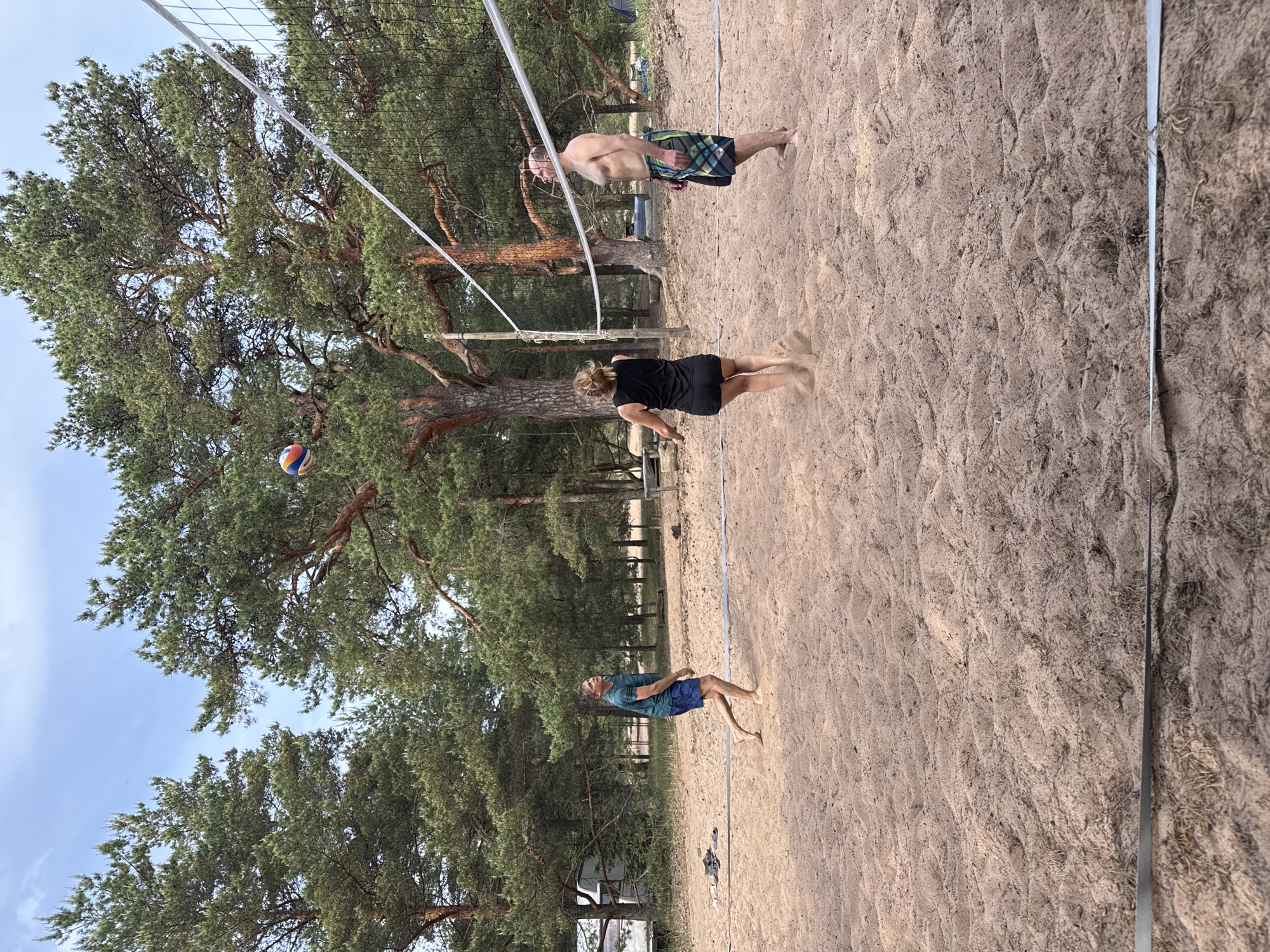 The image captures three individuals engaged in a game of volleyball on a sandy court in Peski, Russia, surrounded by a dense pine forest under a partially cloudy sky.
On the left side of the court, a barefoot man with fair skin, wearing a dark green t-shirt and blue patterned shorts, stands with his body angled towards the right, looking upwards at the volleyball. His left leg is bent, and his right leg is extended backward, indicating movement or readiness. A pair of black sandals lies on the sand near his position.
In the center-right, a barefoot woman with fair skin and light brown hair tied back, wearing a black tank top and black shorts, is positioned with her back partially to the camera. Her arms are raised, and sand is visibly displaced around her feet, suggesting she has just made contact with or is preparing to hit the volleyball. She is also looking upwards.
On the far right, a barefoot, shirtless man with fair skin, wearing multi-colored plaid swim shorts, stands facing towards the left side of the court, observing the airborne volleyball.
A multi-colored volleyball, featuring blue, yellow, red, and white panels, is suspended in the air above a white-banded volleyball net with black mesh. The net is stretched across the court, supported by poles. The playing surface consists of light brown sand, showing undulations and disturbed patterns from player movement. White boundary lines define the court. The background is dominated by numerous tall pine trees with dark green needles and reddish-brown trunks. A white recreational vehicle is partially visible among the trees in the far left background, and faint wooden structures are discernible further back on the right.