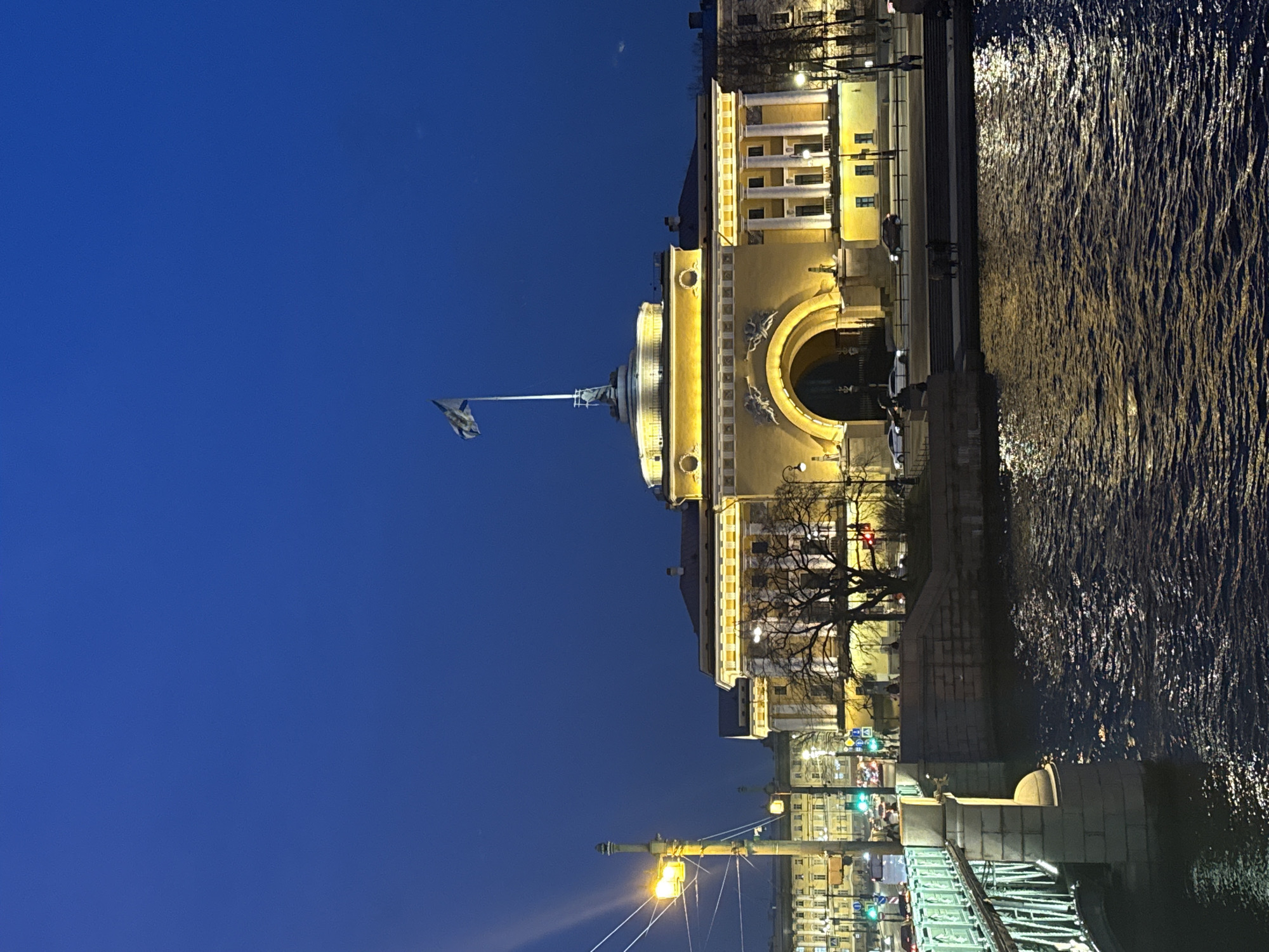 Here's a description of the image:
The image depicts a building in Saint Petersburg, Russia, at night. The building is illuminated with yellow light, featuring classical architecture with a large archway. A flag is atop the building.
To the right of the building is a dark body of water, presumably a river or canal, reflecting some of the light. Below, a bridge is partially visible, with lit street lamps and other buildings in the distance, showing streetlights.
The sky is a deep blue, suggesting it's either dusk or early evening. There are few visible details other than the building, the water, and the sky. The image is rotated.
