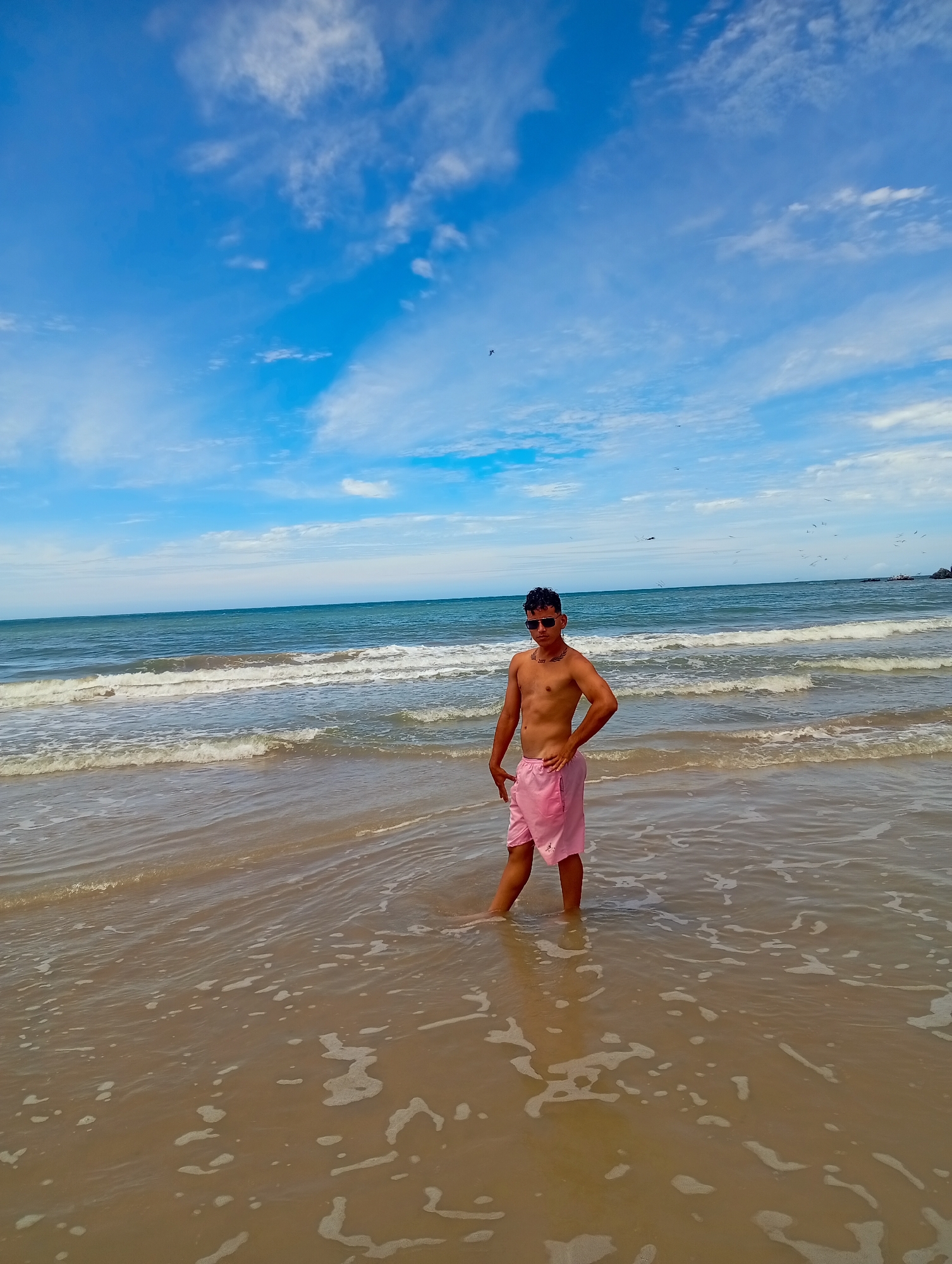 A medium shot captures a person standing in shallow water at a beach, likely on the shores of Altagracia, Venezuela. The individual, who is of medium build, is fair-skinned and wearing sunglasses and pink shorts, appears to be shirtless. He is positioned with one hand on his hip. The water is murky tan with foam on its surface, extending into the frame. Behind the individual, waves are visible, and the ocean stretches towards the horizon where it meets a partly cloudy sky. The sky is blue with scattered white clouds.