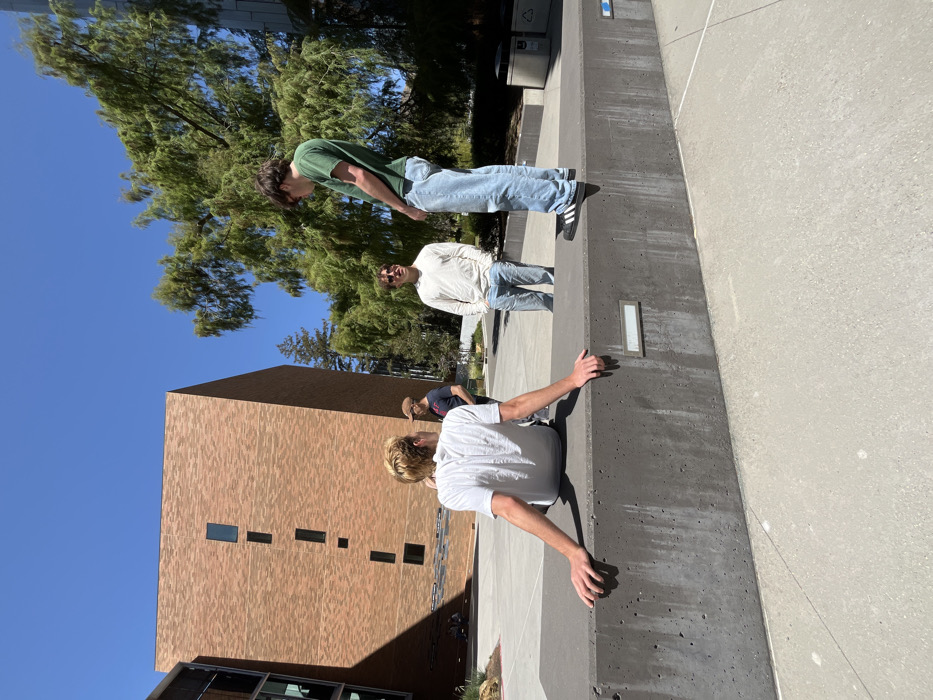 The image captures a scene on the campus of Cal Poly San Luis Obispo. A large, concrete wall is in the foreground with a young man sitting on its edge. He has his right leg crossed over his left, and his hands are on the wall behind him, one on each side of his body. Two men stand on the ground in front of the wall, and a fourth man is behind them, facing the wall. The man in the foreground is wearing a white shirt, and the man to his immediate right is wearing a blue shirt. Both men are looking at the camera. The man furthest from the camera is wearing a white shirt and is standing in front of a tall brick building. The ground is gray concrete, and the sky is blue with white clouds. There is a large green tree in the background. The scene is outdoors and sunny. The building is brick and is part of the Cal Poly San Luis Obispo campus. The coordinates (latitude: 35.302, longitude: -120.661) pinpoint the location as the area near the Cal Poly San Luis Obispo campus.