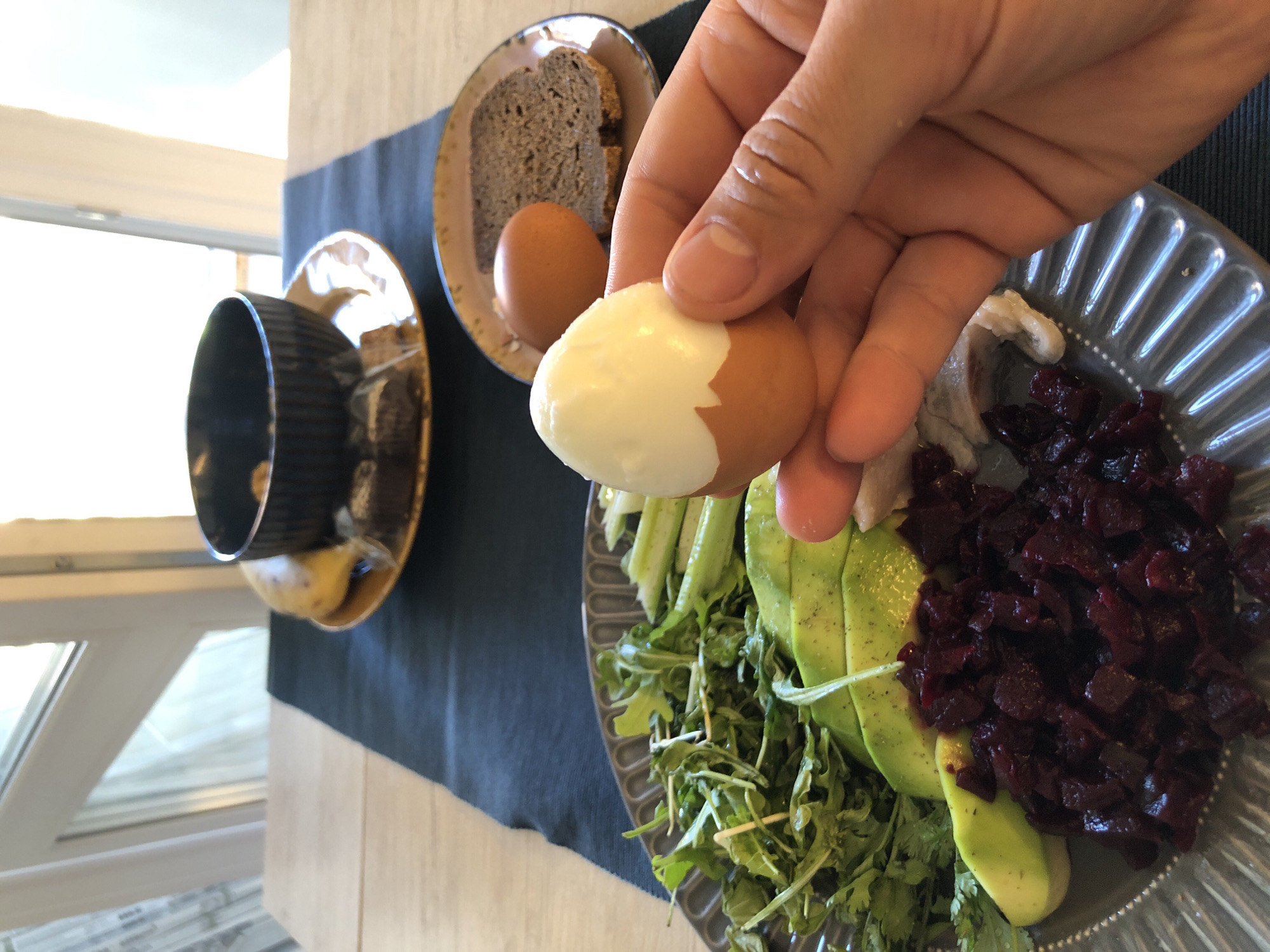 The image captures a first-person perspective of a daytime meal served on a wooden table with a dark blue table runner in Saint Petersburg, Russia. In the immediate foreground, a human hand holds a brown-shelled hard-boiled egg, partially peeled to reveal the white. On a large, grey, fluted plate to the bottom left, a selection of food is arranged: fresh green leaves (likely arugula), several slices of green avocado sprinkled with seasoning, and a pile of diced dark red beetroot. A small portion of white, possibly pickled, fish is also visible near the beetroot. To the right, on a smaller brown plate, rests another whole brown egg and a slice of dark, dense bread. In the upper center, a golden-brown saucer holds a black bowl, possibly containing nuts or granola, and a partially visible yellow banana. A small, dark, packaged item, resembling a brownie, is also on this saucer. The background features a brightly lit window with white frames, indicating an interior setting with ample natural light.