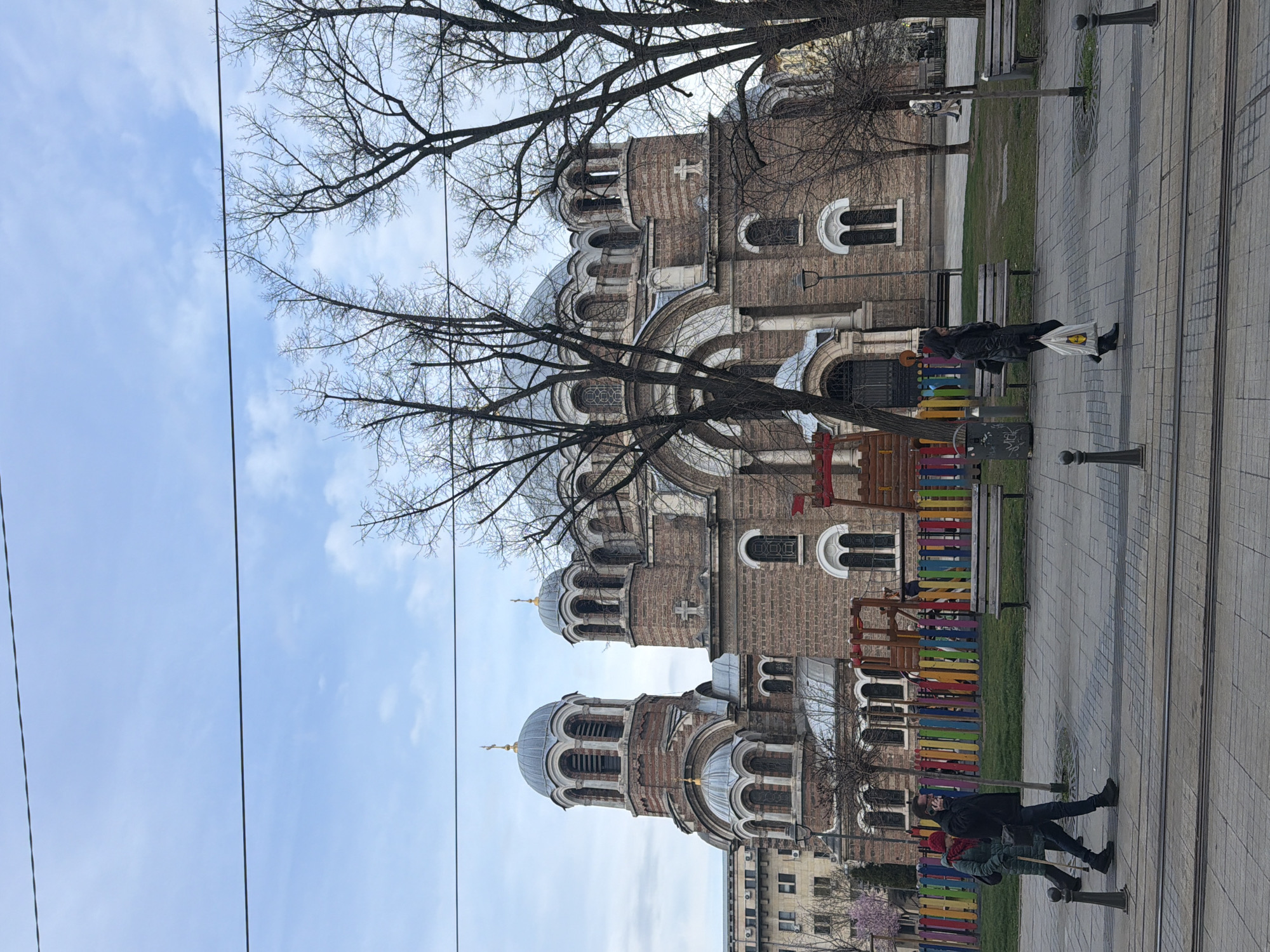 The image shows the St. Nedelya Church in Sofia, Bulgaria, on a bright day with a blue sky and bare tree branches. The church is a brick building with multiple domes and towers. A pedestrian walkway runs alongside the church, with a colorful fence and benches. Two people are walking on the walkway; one is carrying a plastic bag. Power lines are visible in the sky.