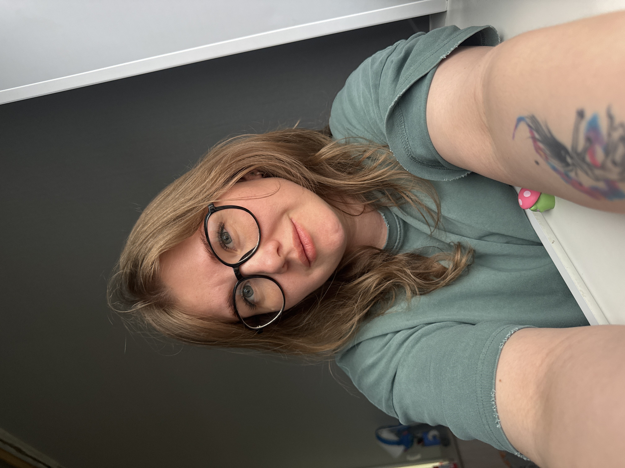 A young woman with light brown hair and glasses looks into the camera, tilted slightly. She wears a green t-shirt. A colorful tattoo is visible on her upper arm. In the background, there is a gray wall and a white ledge. A small mushroom-shaped object sits near her arm. No information in the image indicates the location is Ферма, Russia.