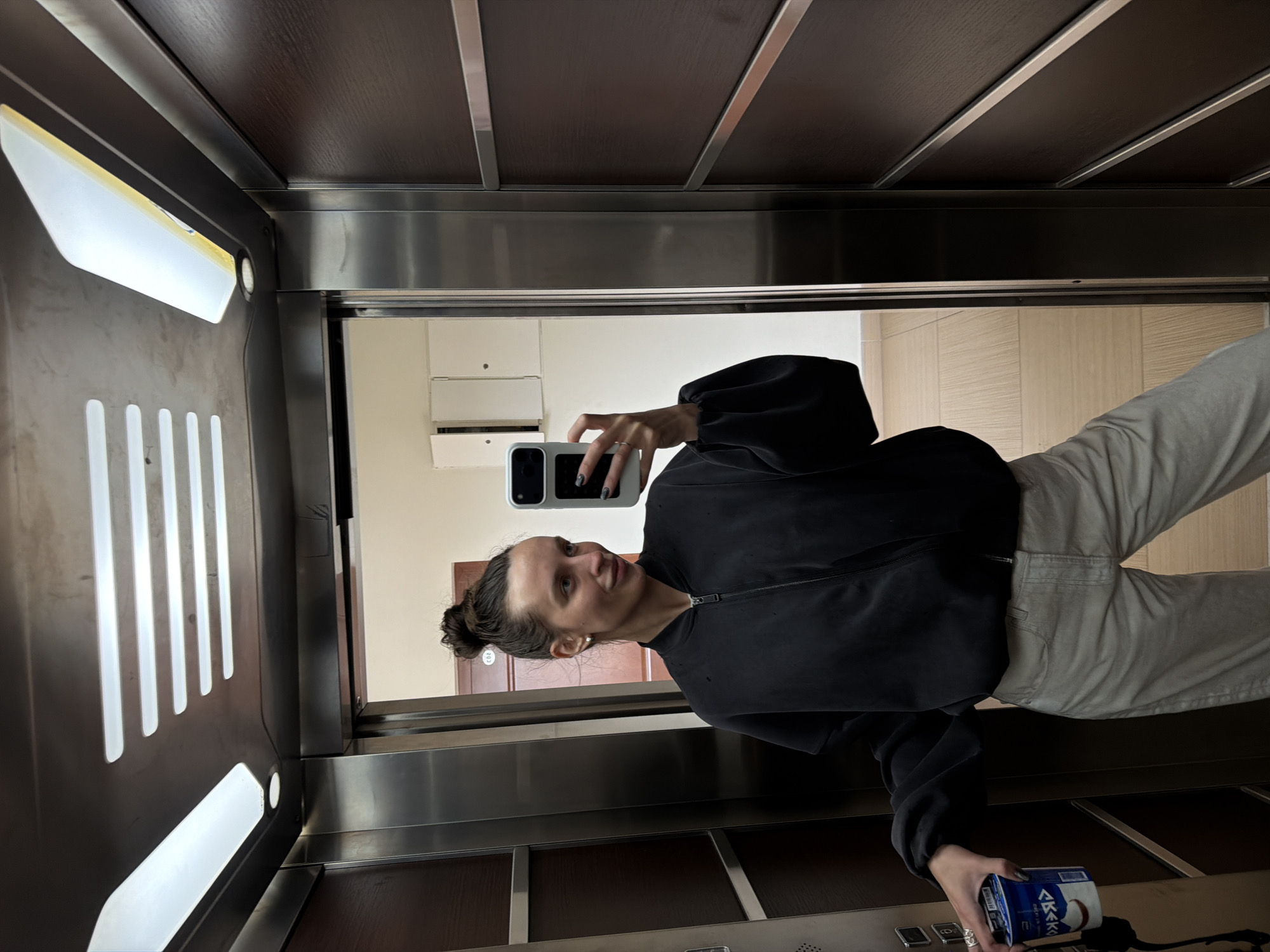 The image, set in Budva, Montenegro, captures a young woman taking a mirror selfie inside an elevator. She is positioned towards the center-right, looking at her reflection with a slight, casual smile. Her dark hair is styled in a bun, and she wears small stud earrings. She is dressed in a black zipped jacket or sweatshirt and light-colored (possibly beige or light grey) pants.
In her raised left hand, she holds a light-colored smartphone with a prominent dark camera module, seemingly capturing the photo. Her fingernails are painted a dark shade. Her right hand, resting lower, holds a blue and white can, possibly a beverage. Cyrillic text and what appears to be "COCONUT" are visible on the can.
The elevator interior features mirrored walls, reflecting the woman and the environment outside the elevator doors. The ceiling has dark panels interspersed with light-colored strips. Bright, angular light fixtures illuminate the space. Through the reflective surface or open doors, a hallway or room with light-toned walls and a dark wooden door, bearing the reflected number "202", can be seen. Part of an air conditioning unit is also visible in the reflection. An elevator control panel is present at the bottom right of the image. The overall scene suggests a casual, everyday moment captured indoors.