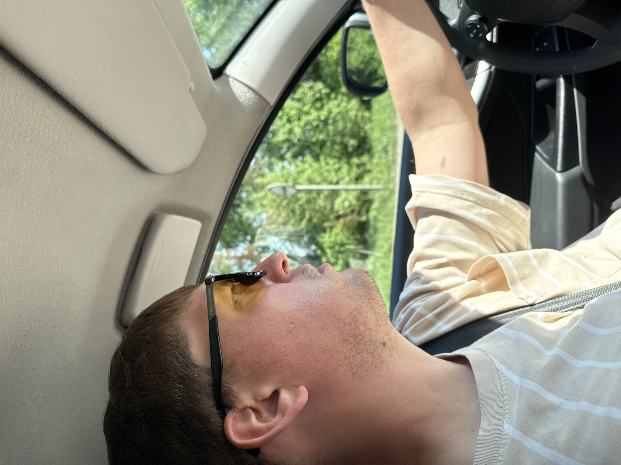A fair-skinned male with short brown hair and stubble lies reclined in the driver's seat of a vehicle. He is wearing sunglasses with yellow lenses and a striped shirt. His right arm is raised towards the steering wheel. The scene is set inside a car, visible through the windows are green trees and what appears to be a street light. The location is assumed to be Dvigatel', Russia, given the prompt's instructions, however, there is no evidence to support this statement.