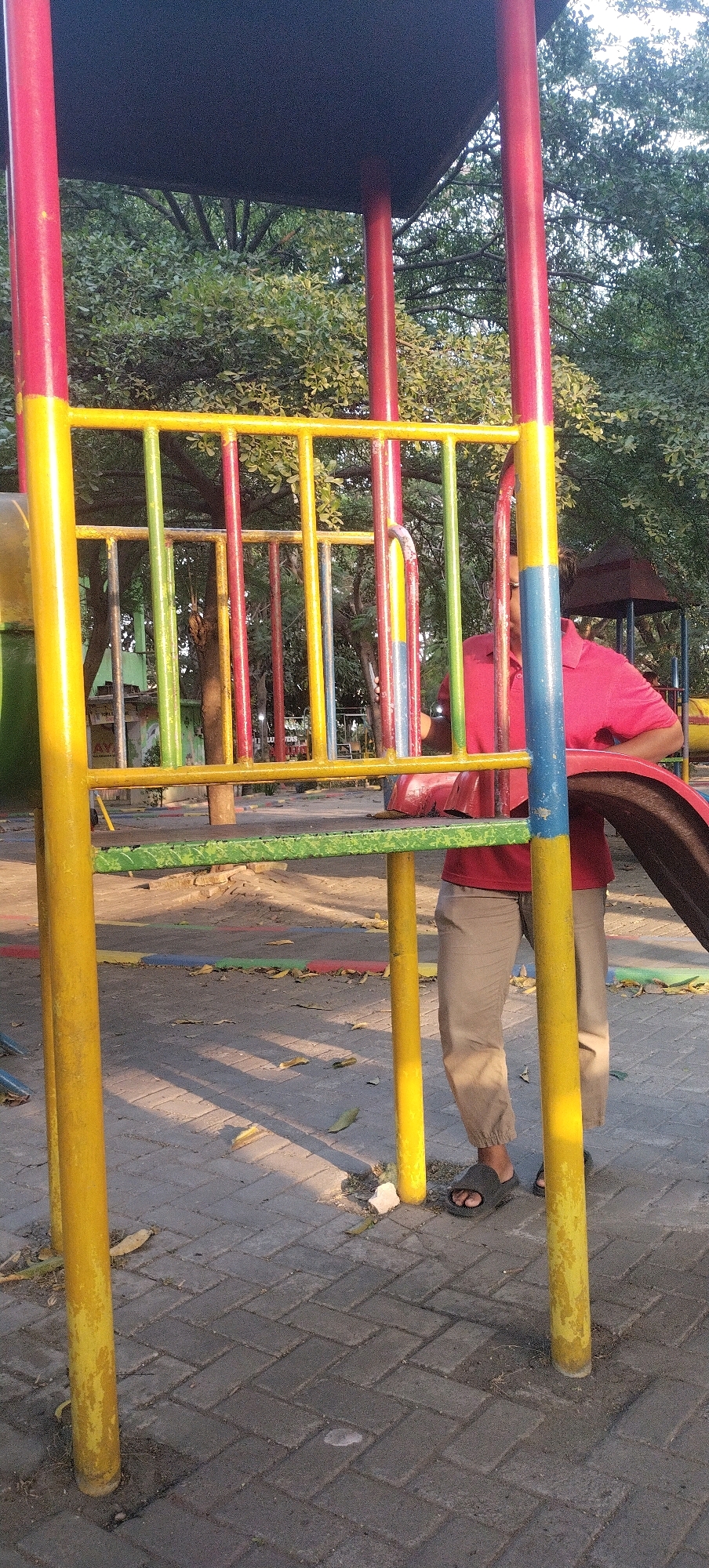 The image shows a playground with a person standing behind a colorful structure. The playground equipment is made of metal poles painted in red, yellow, green, and blue, with a yellow horizontal bar across the front. The ground is paved with brick-like stones, and there are some fallen leaves on the ground. In the background, there are trees and a building with a brown roof. The person is wearing a red shirt and tan pants, and is partially obscured by the playground equipment. The lighting suggests it is daytime, with shadows cast on the ground. The location is identified as Gayam, Indonesia.