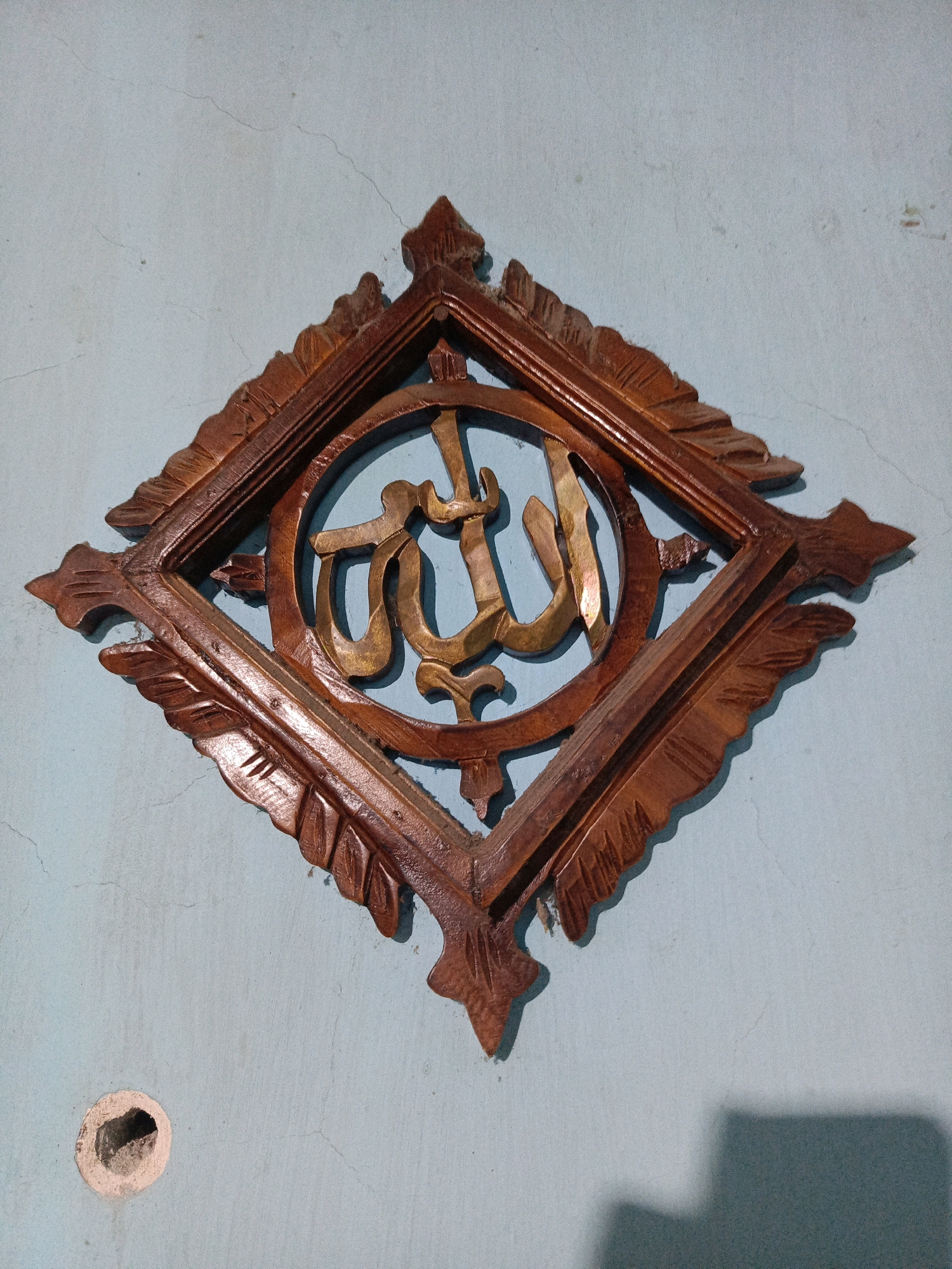 The image shows a close-up, eye-level, medium shot of a rhombus-shaped wooden wall decoration hanging on a light blue, textured wall. The decoration features carved leaf motifs on its outer edges and a circular frame in the center, within which is a golden Arabic calligraphy of "Allah". The wall has visible cracks and a small, circular hole. The lighting suggests an indoor setting, possibly a home, with a shadow cast on the lower right side of the wall. The image was taken in Kalang, Indonesia.