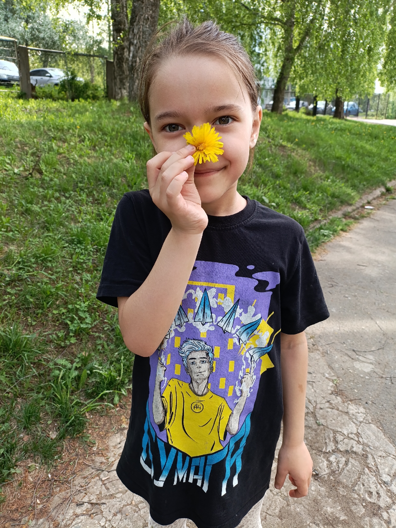 A young girl stands on a concrete path holding a dandelion flower near her nose. She is wearing a black t-shirt with a cartoon graphic featuring a stylized male figure with blue hair and yellow shirt. The background consists of green grass, trees, and a glimpse of parked cars. The location is consistent with a public park or recreational area in the city of Chaikovsky, Russia.