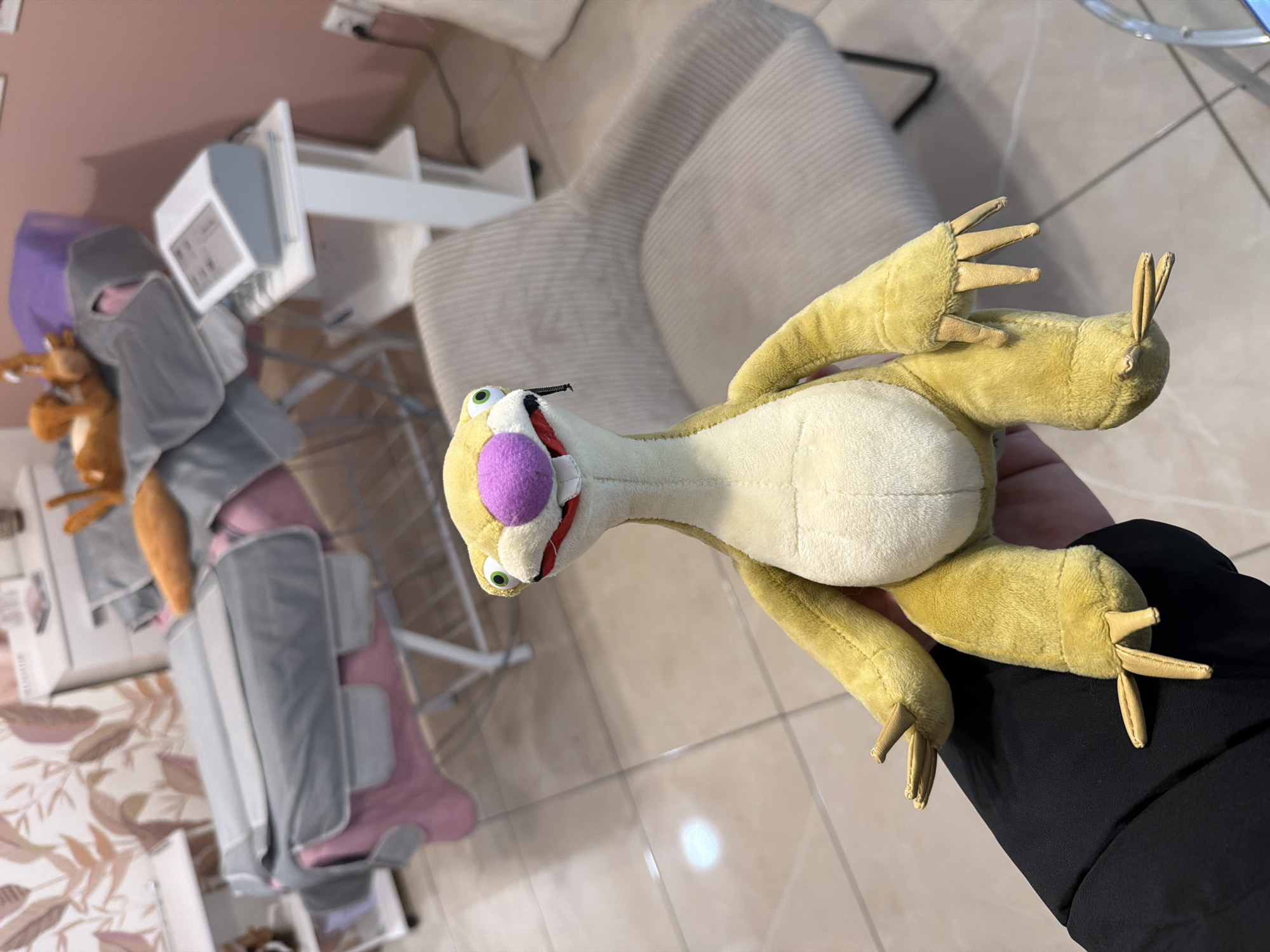 A plush toy sloth with yellow fur, a white belly, and a purple nose is held by a person wearing a black garment. The background includes a chair with a striped pattern, a compression device on a pink surface, and a wooden floor. The location is possibly Burgas, Bulgaria, though this information is not visually verifiable within the image itself.