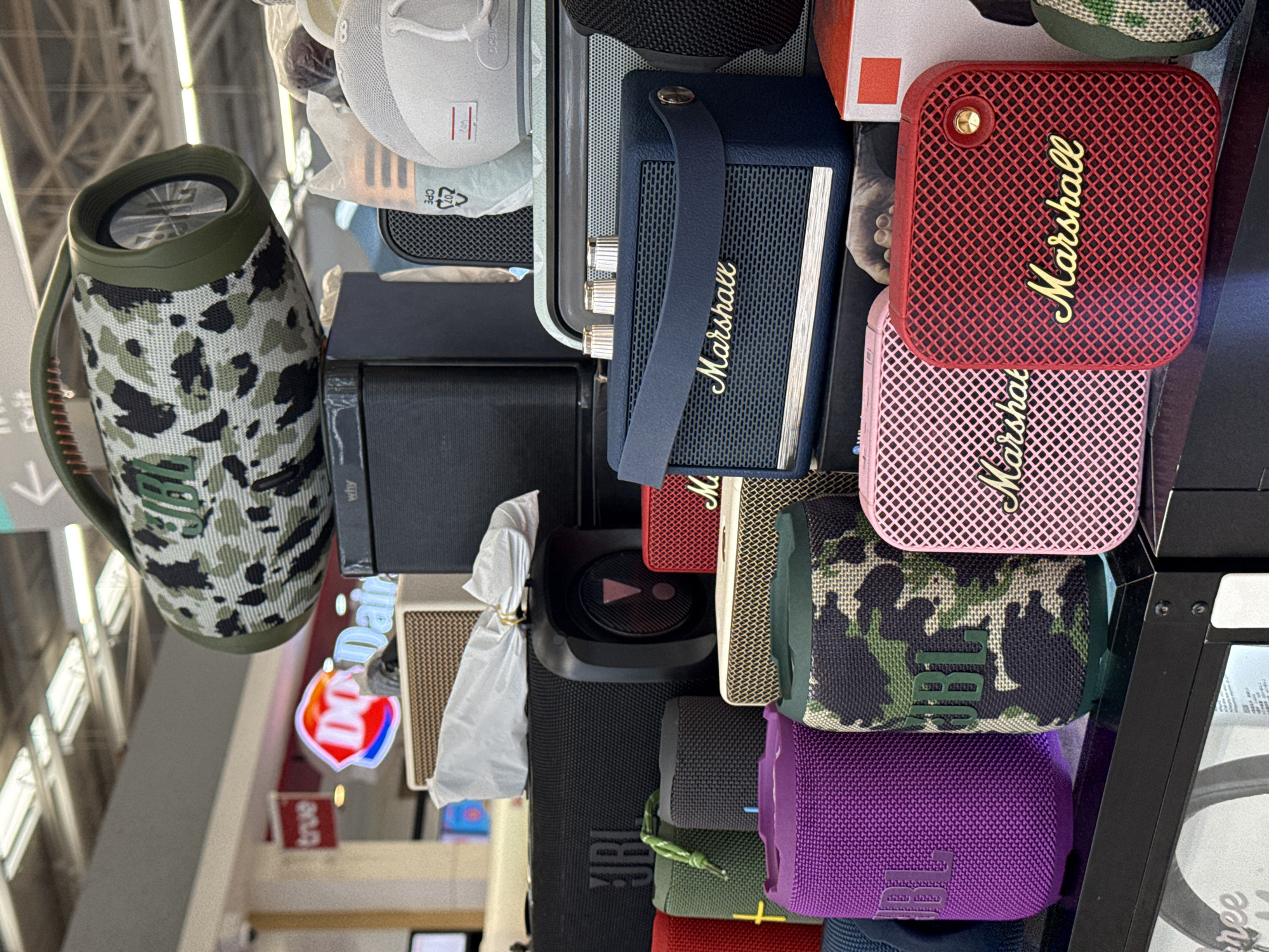 The image depicts a retail display of portable Bluetooth speakers in a store. The foreground features several speakers of various sizes and colors, including purple, camouflage, pink, and red. Brands like JBL and Marshall are visible on some of the devices. In the background, a large camouflage speaker with the JBL logo is prominently displayed. Other elements in the background include store signage, such as a red "true" sign and a Dairy Queen logo, suggesting the location is a shopping mall or electronics store. The overall environment appears to be indoors, with artificial lighting. The image does not contain any people or explicit actions. The scene is static and focuses on the product presentation.