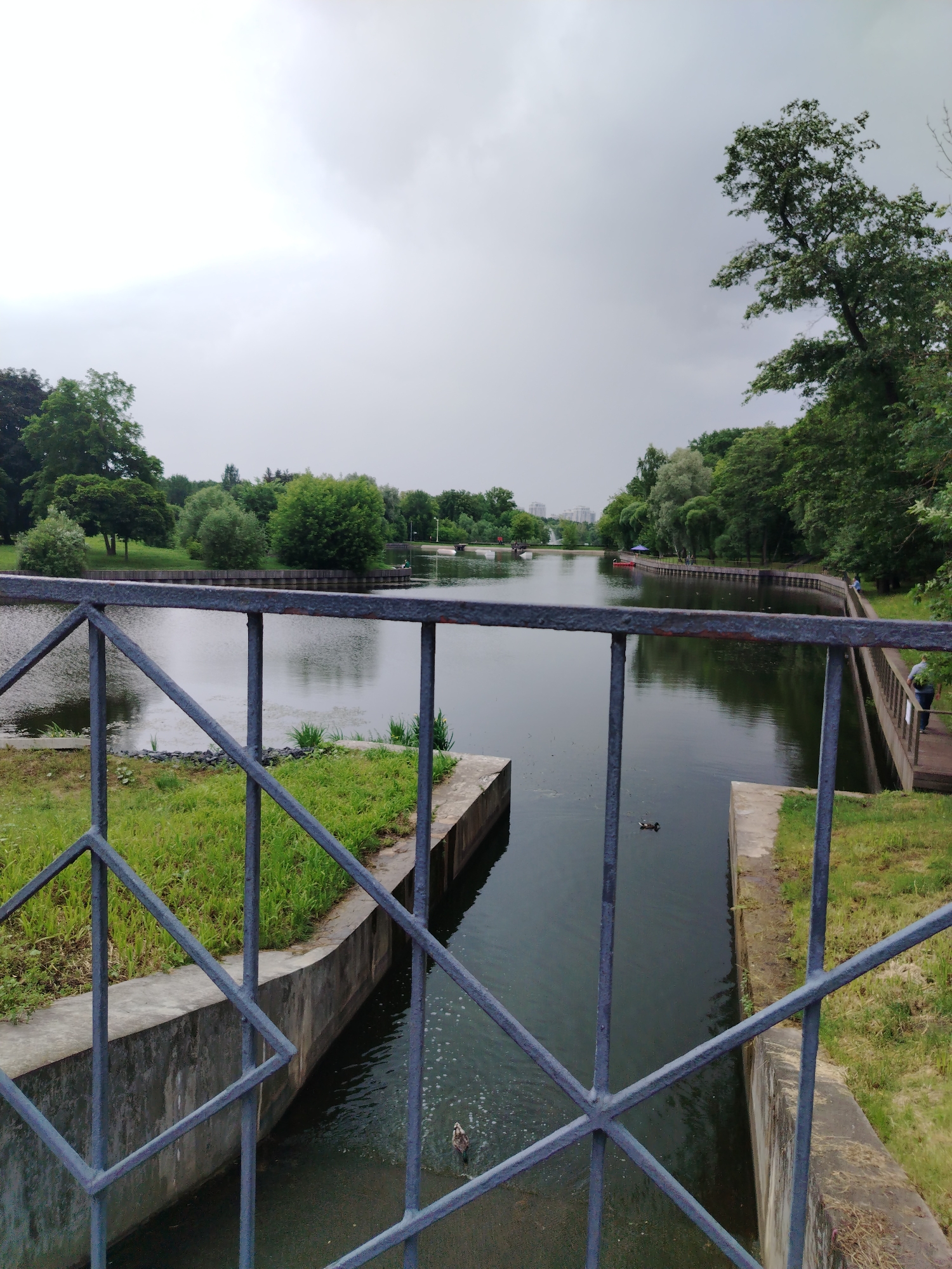 This is a vertical, eye-level photograph of a canal or river in Moscow, Russia, under an overcast sky. The foreground features a dark grey metal railing with a diamond pattern, obscuring the bottom portion of the scene. Beyond the railing, a concrete embankment lines the left side, leading to a body of dark water. Green grass grows along the top of the embankment.
The water stretches into the distance, reflecting the cloudy sky. On the right side, a wooden walkway or pier runs along another concrete embankment, with several people visible. A person in a blue shirt is walking away from the camera on this walkway. Further down the canal, small boats are present, some appearing to be red. Trees and lush green foliage line both banks of the waterway, creating a park-like setting. In the far distance, buildings are visible, suggesting an urban environment integrated with nature. The overall mood is serene and slightly subdued due to the cloudy weather, perhaps indicating late morning or early afternoon. A duck is visible swimming in the water in the middle ground. There is no visible text in the image.