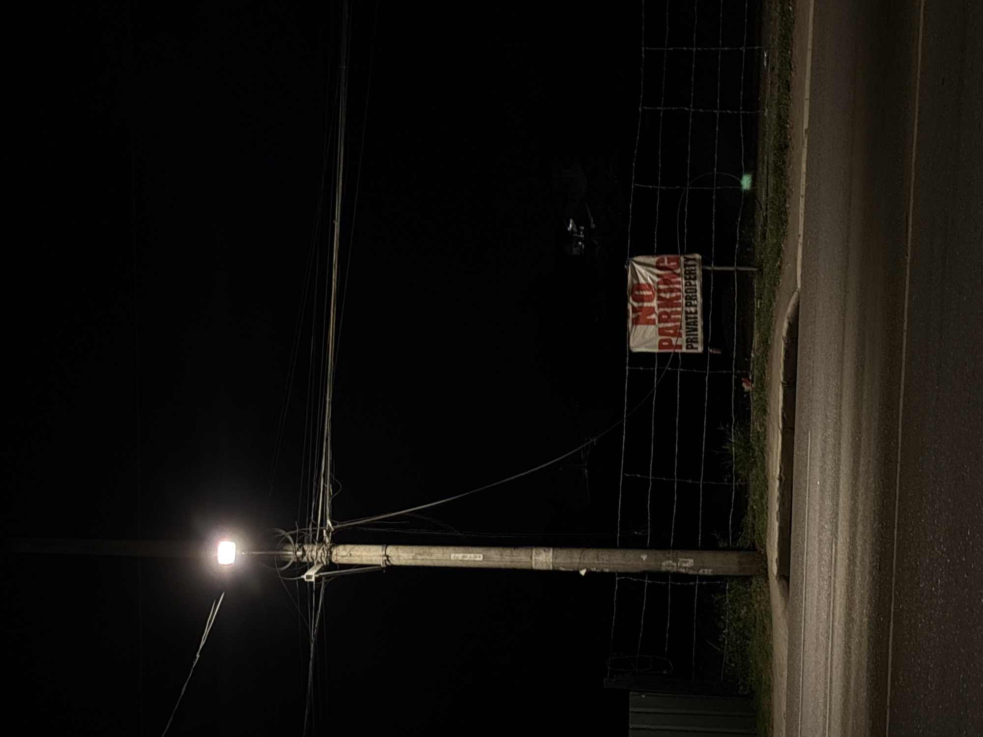 The image captures a static, nighttime street scene in Davao City, Philippines. A prominent, light-colored utility pole stands on the left side of the frame, supporting an active street light fixture that emits a bright, warm white glow, illuminating the immediate vicinity. Numerous intertwined electrical and communication wires are bundled and strung from the pole, extending horizontally across the mid-upper section of the scene, with some individual wires visibly slack.
In the foreground, a dark asphalt road surface displays two faint, light-colored parallel lines. This road is bordered by a concrete curb, behind which lies a narrow strip of unkempt ground with sparse patches of grass and dirt. Further back, a multi-stranded wire fence, possibly barbed or plain, stretches horizontally across the middle ground, demarcating a property boundary. Attached to this fence, to the right of the utility pole, is a white rectangular sign with bold red text clearly stating "NO PARKING" above "PRIVATE PROPERTY".
The area beyond the fence and in the upper portions of the image is predominantly dark, obscuring details, though faint outlines of distant structures or vegetation can be indistinctly observed in the deeper background. A section of a dark, corrugated metal fence is visible on the far left, behind the utility pole. A subtle green light or reflection appears on the ground behind the wire fence, to the right of the "No Parking" sign. No individuals or active subjects are present in the depicted environment.