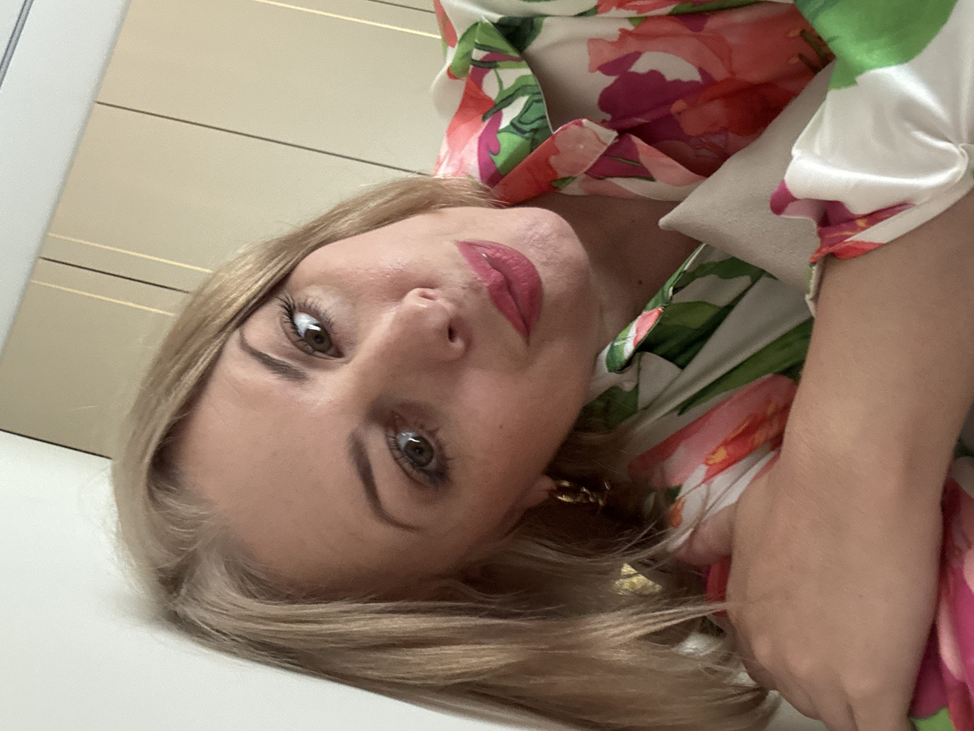 This is a close-up, portrait-style media file of a woman. She has blonde hair, is wearing a white top with a colorful floral print in pink and green, and has bold pink lipstick on. Her eyes are open and looking towards the camera. She is wearing a gold hoop earring. The lighting suggests it could be daytime indoors, possibly in a well-lit room. The background appears to be a neutral-colored wall with horizontal lines. The image is taken at an angle, with the woman's head tilted. There is no visible text in the image. The setting cues suggest a modern indoor environment, and the woman's expression is neutral. There are no specific indications of weather or time of day beyond general indoor lighting.