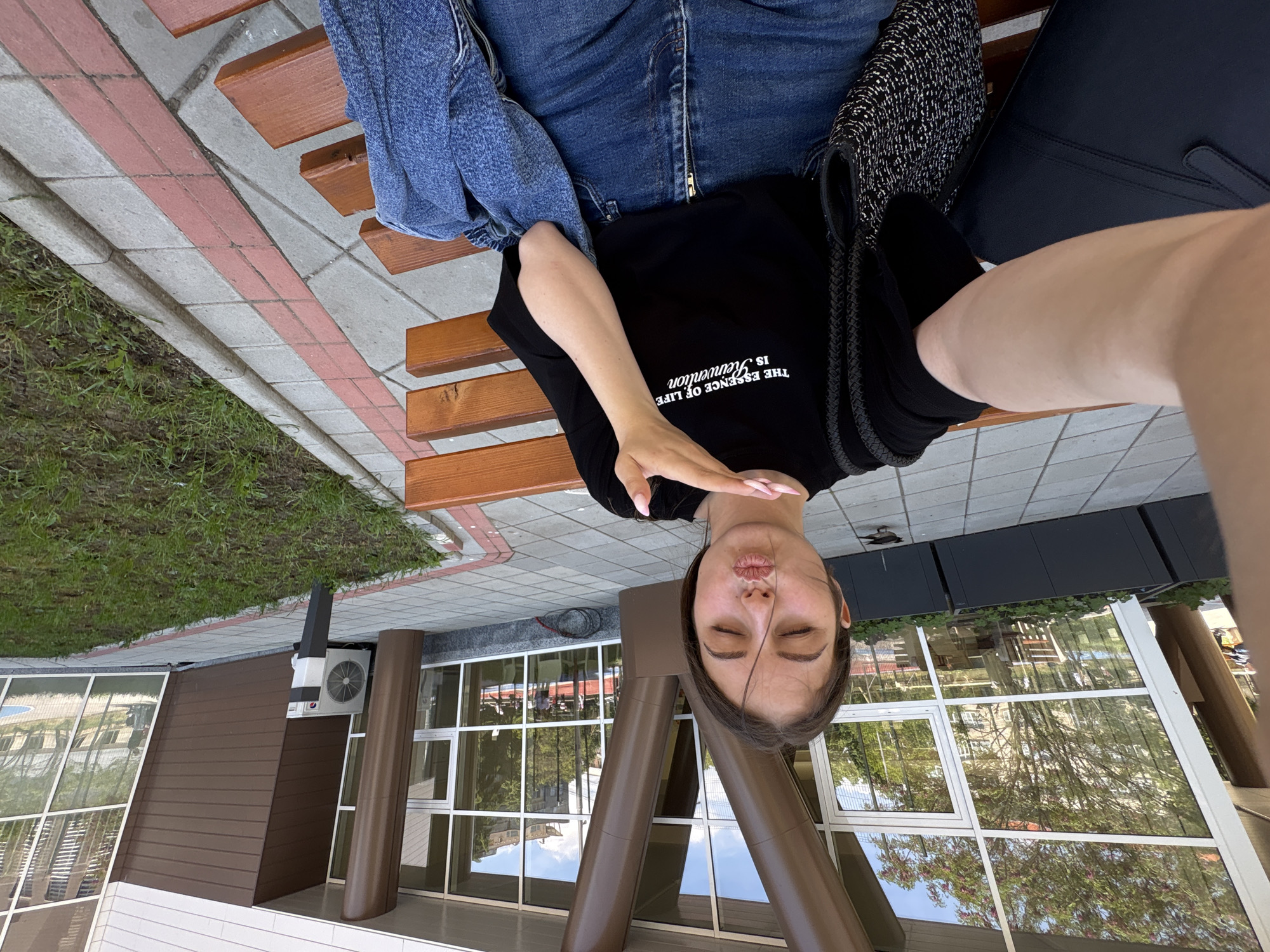 A woman with brown hair is taking a selfie on a bench. She is wearing jeans and a black t-shirt with white text. She is sitting on a wooden bench in front of a building with large windows. The location is Burgas, Bulgaria. There is grass and pavement around the bench and the building. There are bags next to her on the bench.