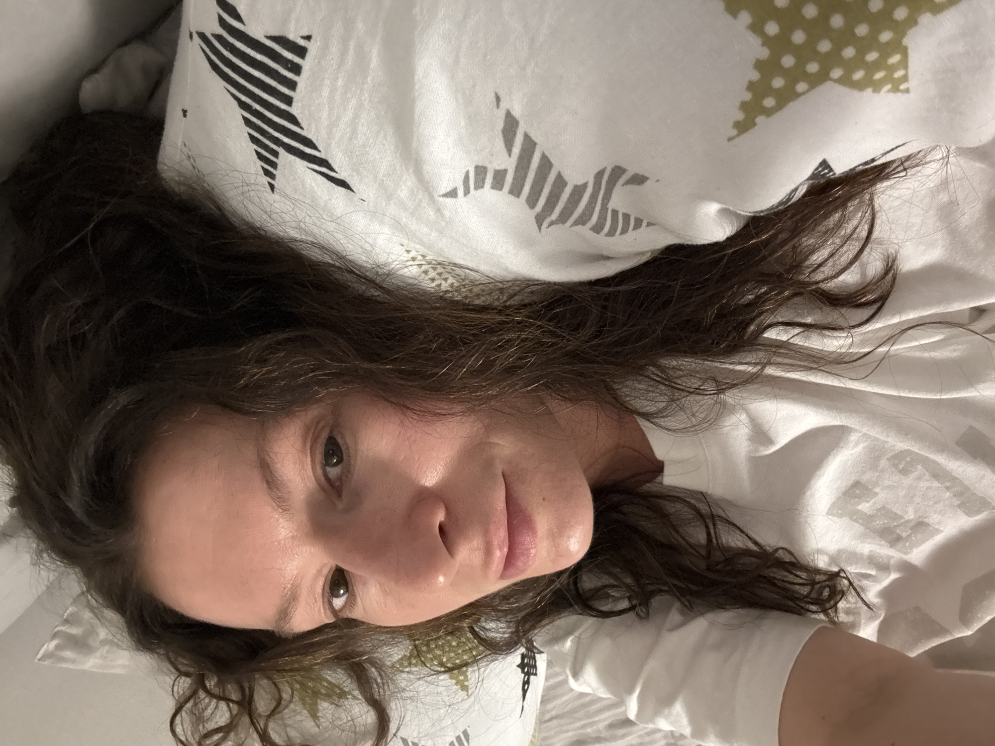 A woman with long, wavy, brown hair is lying down on a bed. She is looking at the camera. Her skin tone is light, and her expression is neutral. She is wearing a white T-shirt. The bedding consists of a white sheet and pillow with star patterns of varying colors. The image appears to be taken indoors, in a bedroom setting. Location in Chaikovsky, Russia is unknown.
