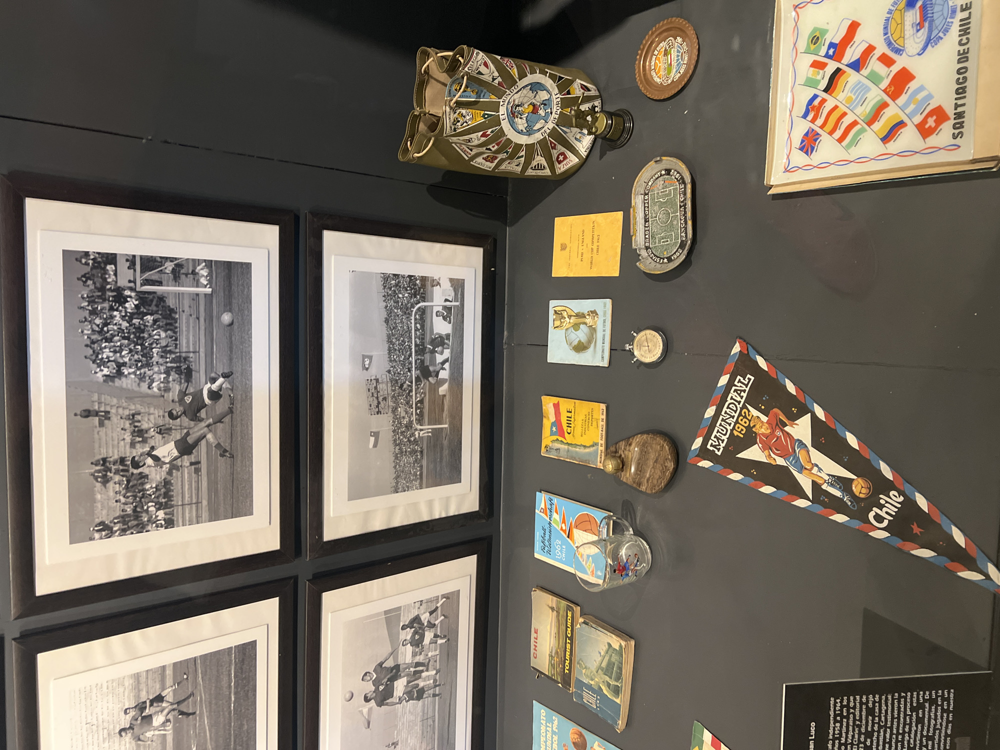 This is a photograph of a wall display, likely in a museum or exhibition, relating to the 1962 FIFA World Cup held in Chile. The display includes black and white photographs of soccer matches, a pennant reading "MUNDIAL 1962 Chile," a bag decorated with flags and labeled "EL MUNDO DEL DEPORTE," commemorative plates and items related to the World Cup, and a glass plaque of flags and writing: "SANTIAGO DE CHILE." There are also several Chilean tourist guides. A descriptive text panel is also displayed in the bottom right corner. The photographs are in dark brown frames. The display surface is a dark matte gray. The location (Arica, Chile) is not evident from the image.