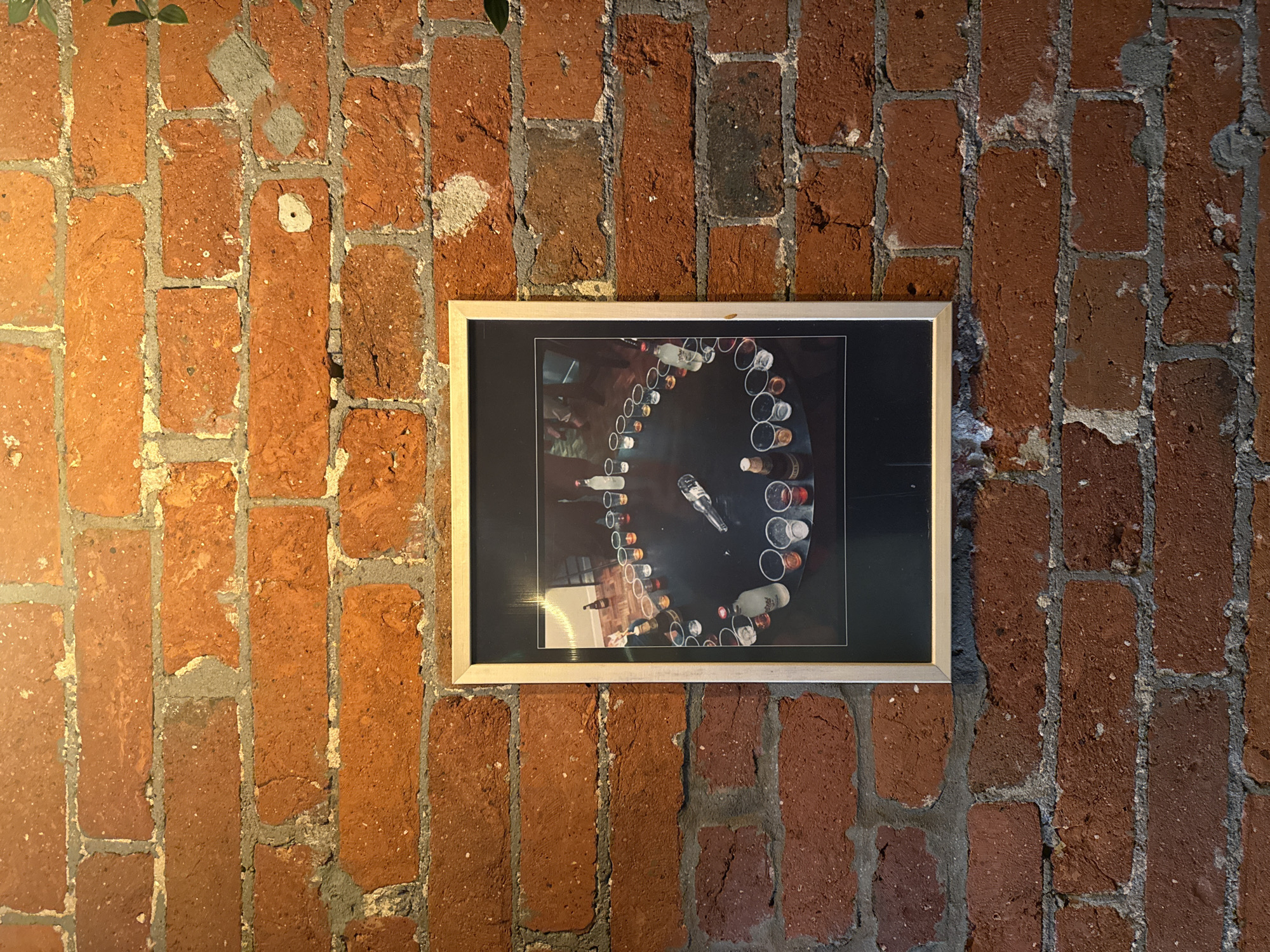 The image shows a framed photograph hanging on a red brick wall. The photograph features a circular arrangement of bottles and glasses on a dark surface. The wall appears to be an interior wall constructed of old bricks with visible mortar. The lighting is somewhat uneven, suggesting ambient light sources. The location in Moscow, Russia is not discernible from the image.