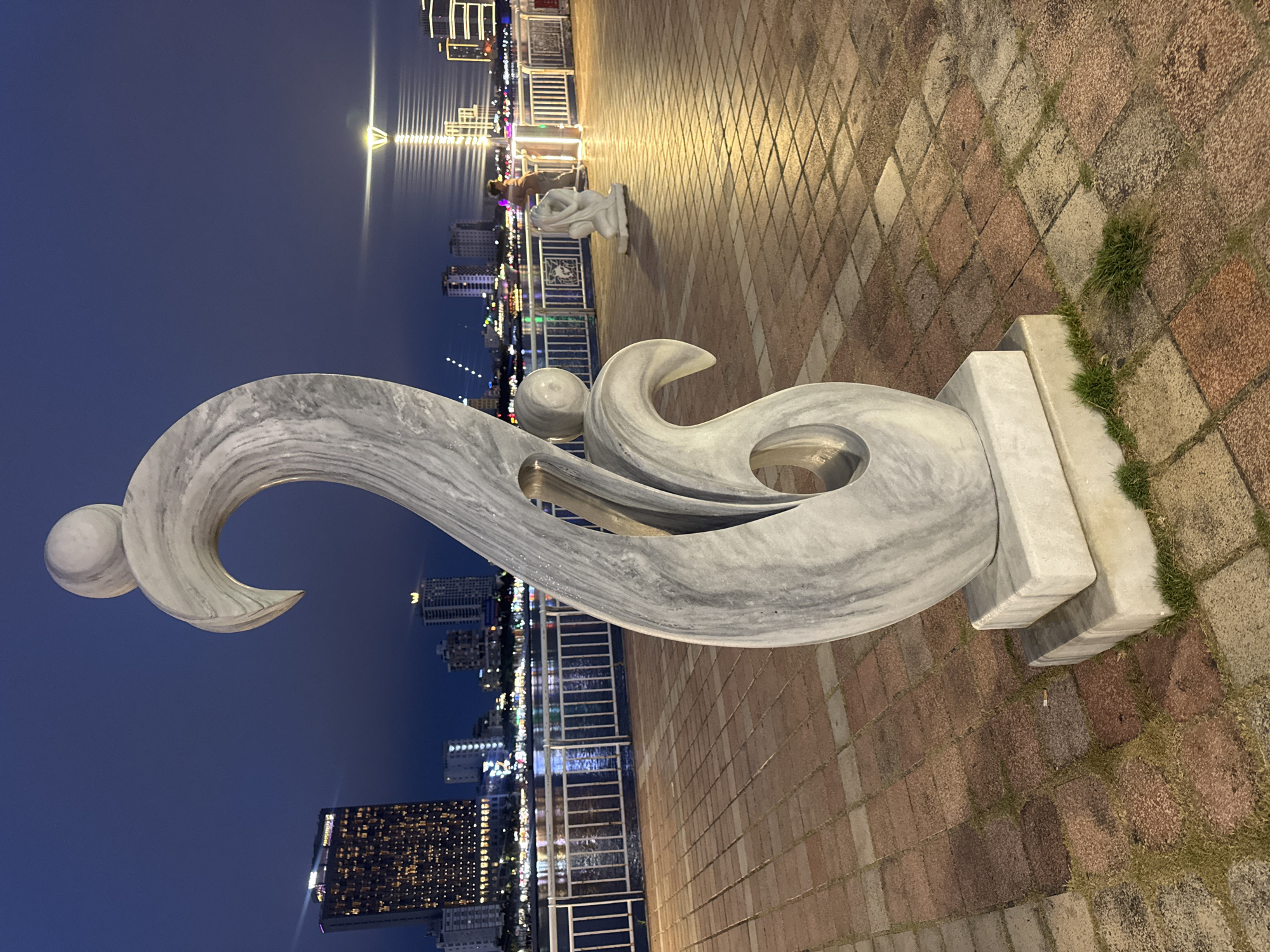 The photograph depicts a marble sculpture on a brick-paved promenade at night. The sculpture is primarily gray and white with swirling patterns and is mounted on a light-colored base. A metal railing runs behind the sculpture. In the background, the cityscape of Đà Nẵng, Vietnam is visible, illuminated with numerous lights. The sky is dark blue. A street light is reflected in the water. Another marble sculpture is seen in the background. The area is well-lit. The tiles of the pavement are arranged in a repeating geometric pattern. Some grass grows around the sculpture's base.