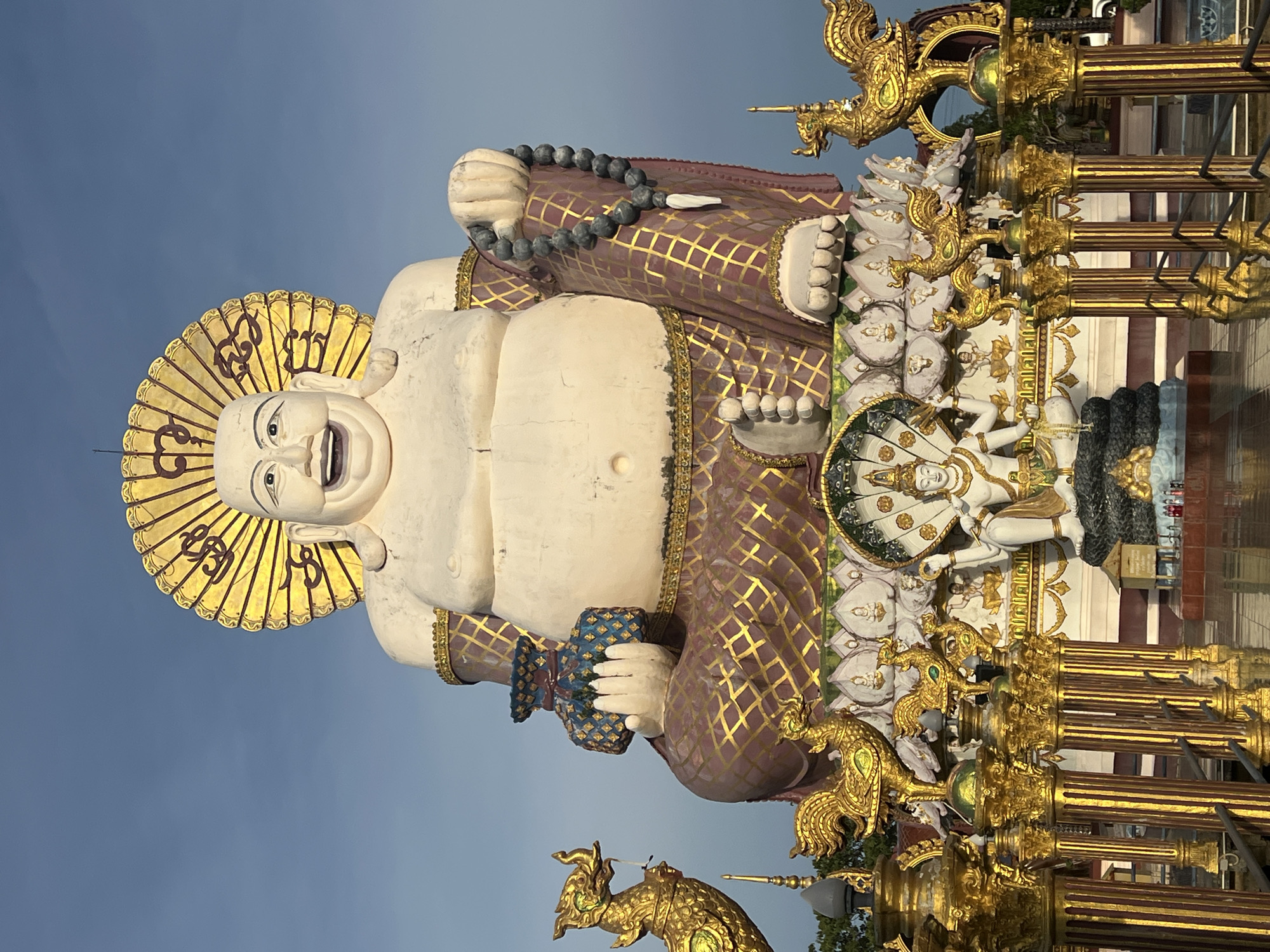 The image captures a full-body, low-angle shot of a monumental statue of a laughing Buddha, known as the Big Buddha of Koh Samui, located at Baan Plai Laem temple on Koh Samui island, Thailand. The statue is predominantly white, with a large, round belly and a joyous, open-mouthed expression. It wears a golden halo behind its head, adorned with swirling patterns. The statue is draped in a purplish robe with intricate gold geometric patterns and gold trim. In its left hand, it holds a blue and gold patterned pouch, while its right hand rests on its lap, holding a string of dark gray prayer beads with a white pendant.
The statue is seated on a multi-tiered, ornate base decorated with lotus motifs and smaller statues. Below the main Buddha statue, there is a central figure in white, seated cross-legged with hands raised, possibly another deity or representation. The entire structure is surrounded by vibrant gold ornamentation, including ornate pillars topped with golden mythical bird figures, resembling phoenixes or similar creatures. The background is a clear, pale blue sky, indicating daylight. The scene is illuminated by the sun, casting a warm glow on the golden elements. The overall impression is one of grandeur and spiritual significance within a tropical setting.