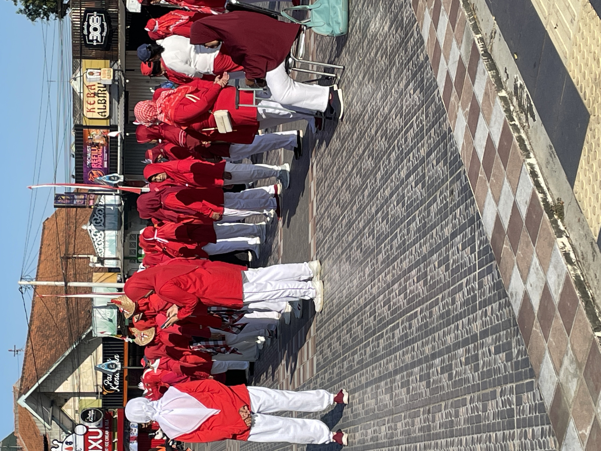 A group of people, primarily women, are gathered outdoors on a paved area, likely in the city of Purworejo, Indonesia, given the context of the image. They are mostly wearing red tops and white pants, with many also wearing headscarves, predominantly red or white. The scene appears to be in front of several buildings with various signs, including "IXU," "Pai Kenan," "SPECIALIST REFILL PARFUM," "KEBA ALBIRU," and "308." Some individuals are standing in rows, while others are interacting or observing. One woman is seated in a chair, wearing a maroon headscarf and white pants, with a light blue bag next to her. The ground is paved with patterned tiles. The lighting suggests it is daytime, with sunlight casting shadows. The overall atmosphere seems to be that of a gathering or event.