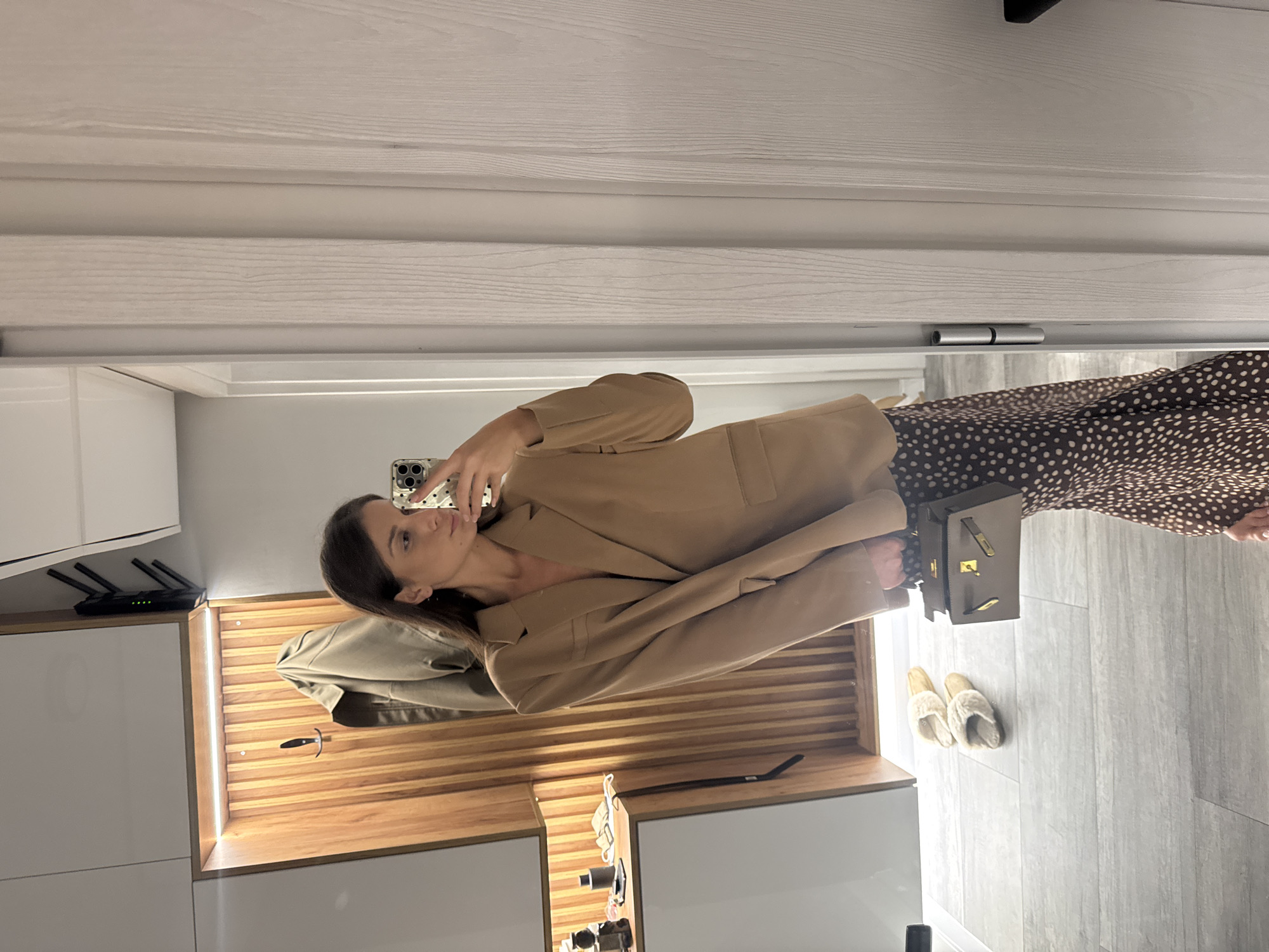 A woman with dark hair is taking a mirror selfie indoors. She is wearing a brown blazer and a brown and white polka-dot dress. She is holding a cell phone in her left hand. The location appears to be a modern apartment or home interior with a wooden accent wall and light grey floors. A handbag and slippers are near her feet. The setting is unlikely to be in Burgas, Bulgaria, without additional context or confirmation.
