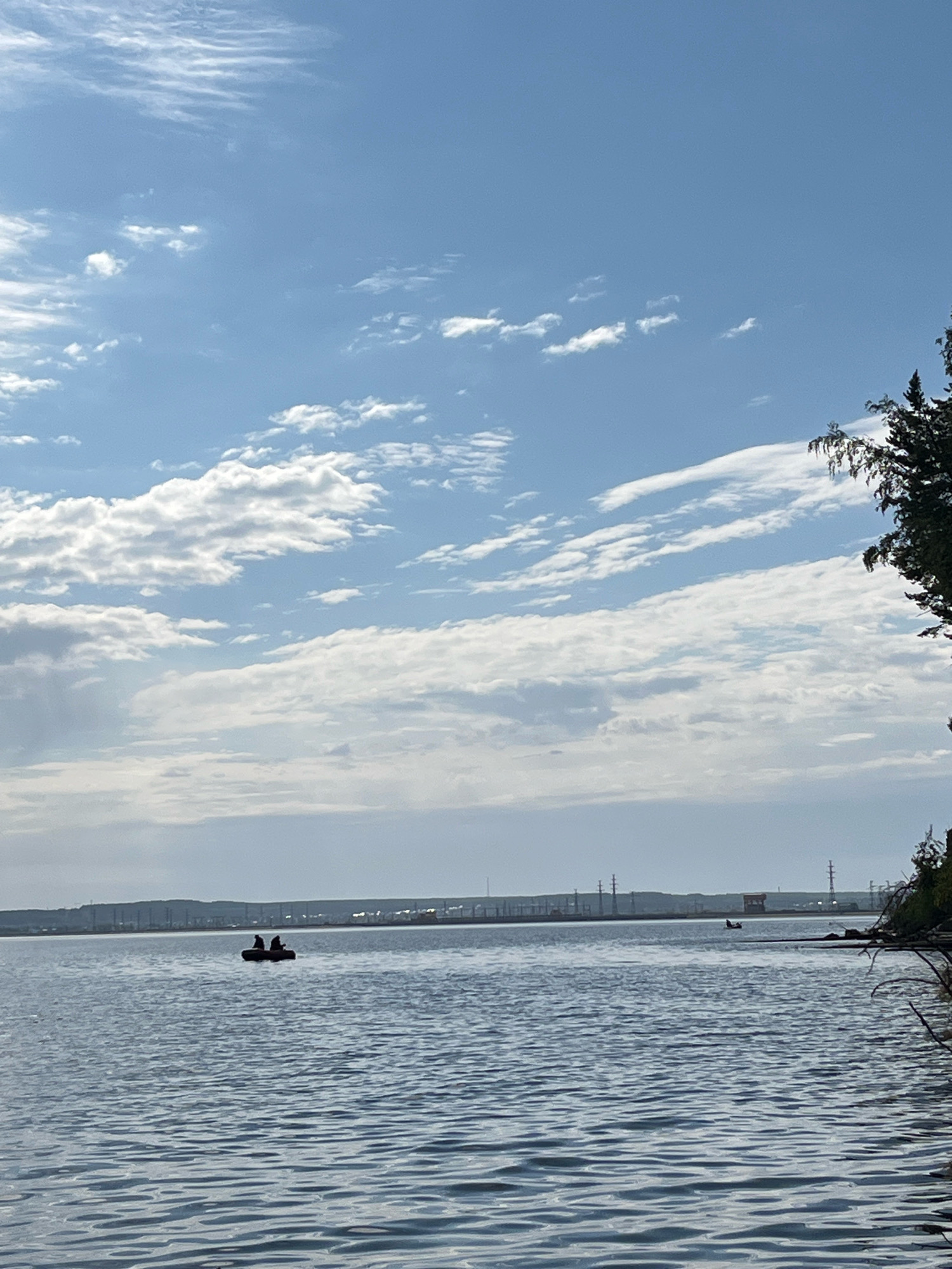 The image depicts a tranquil scene on the Kama River in Chaikovsky, Russia. The sky is a brilliant blue, dotted with scattered white clouds that stretch across the horizon. The water is calm, with gentle ripples reflecting the sky.
In the foreground, the vast expanse of the river occupies most of the frame, with the distant shoreline visible as a faint line on the horizon. The shoreline is characterized by a somewhat industrial landscape, with power lines and structures suggesting human activity.
Further out on the water, a small, dark-colored inflatable boat is visible. Two figures, silhouetted against the bright sky, are seated in the boat, seemingly engaged in fishing or enjoying a leisurely row. The boat is positioned in the middle ground of the image, adding a sense of scale to the expansive river.
To the right of the frame, a cluster of trees with lush green foliage stands on the bank, framing the scene and adding a touch of natural beauty. The overall impression is one of peace and serenity, with the gentle movement of the water and the distant figures in the boat contributing to a sense of calm.