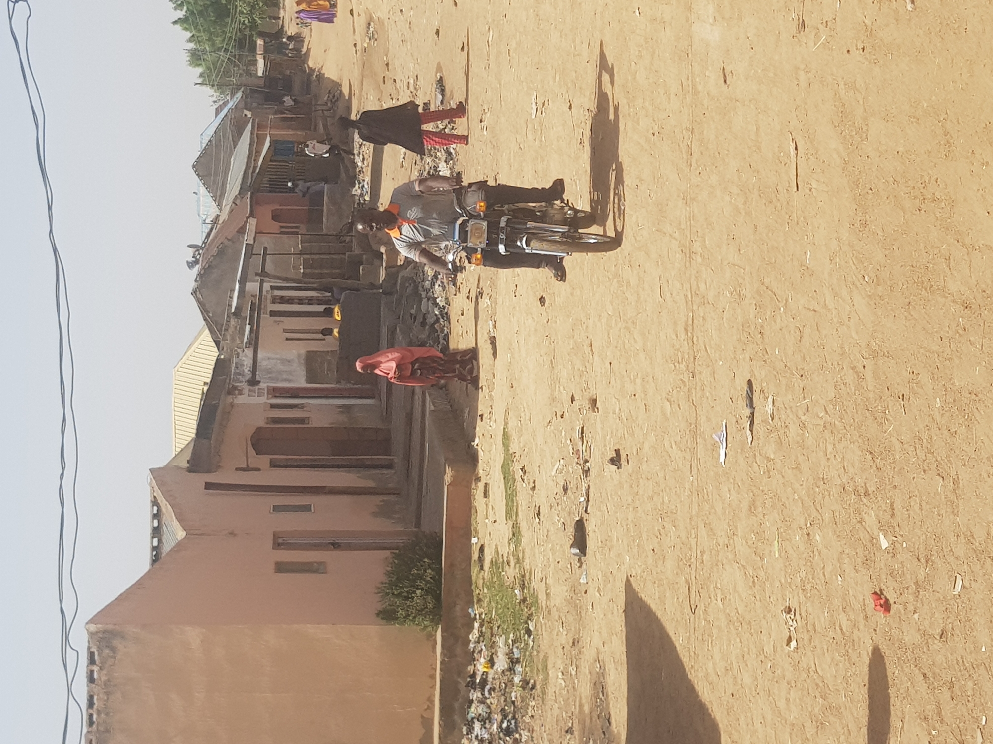 The image depicts a street scene in Dabawa, Nigeria. The dirt road is bordered by buildings constructed from mud brick, with some having corrugated metal roofs. Litter and debris are scattered along the road. A man on a motorcycle is traveling along the road, and other individuals are walking nearby, including a person in red clothing. The environment appears arid and sunny.