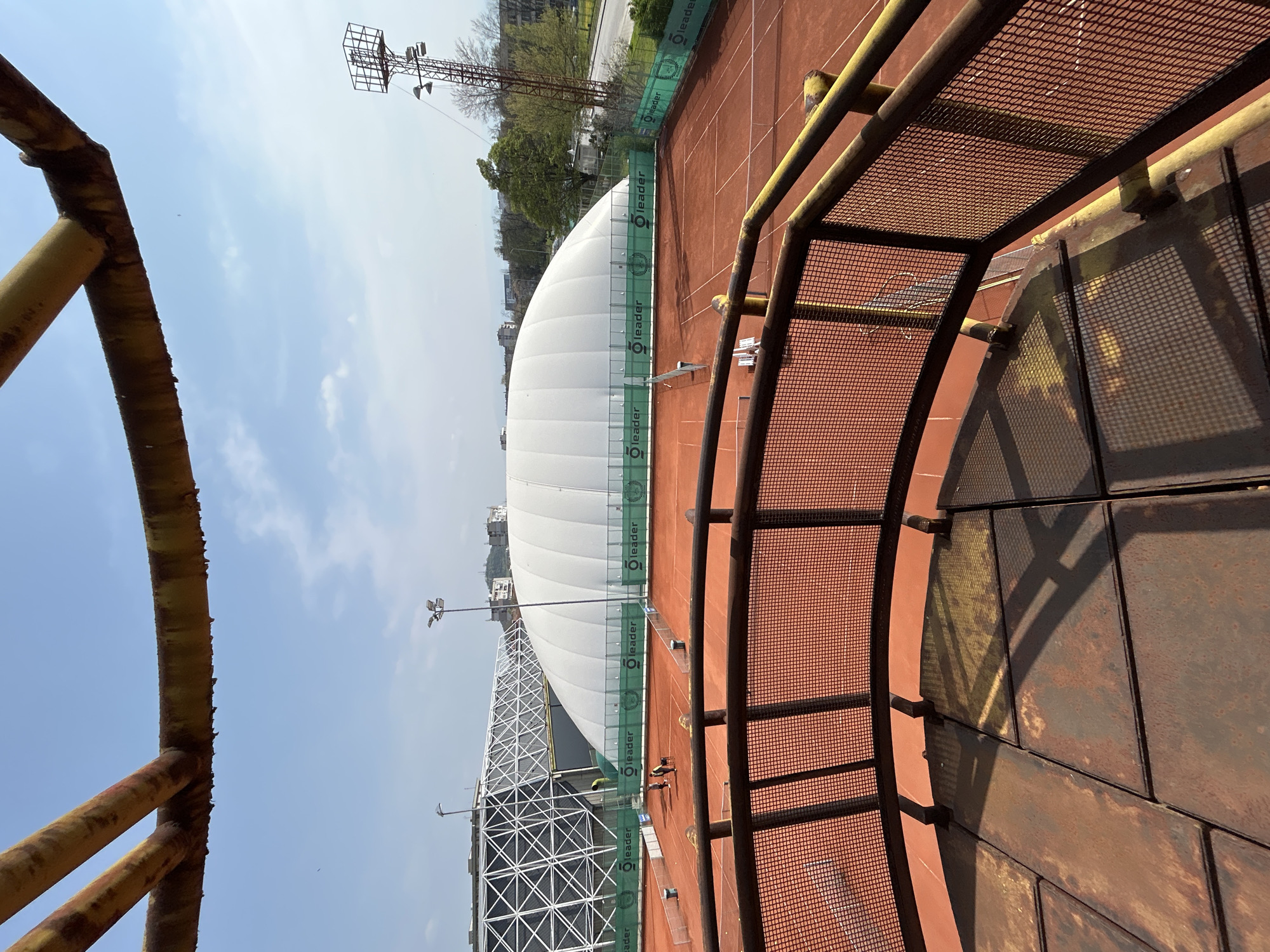 The image shows a tennis court complex with a large, white, dome-shaped structure covering one of the courts, likely an air-supported structure for year-round play. The court surface is red clay. A tall metal light tower is visible. The view is from an elevated platform or structure, featuring rusty metal grating and railings. Two people are visible on the tennis court. The scene is situated in Plovdiv, Bulgaria, as indicated by the "leader" branding on the court dividers, which is associated with tennis facilities in Plovdiv.
