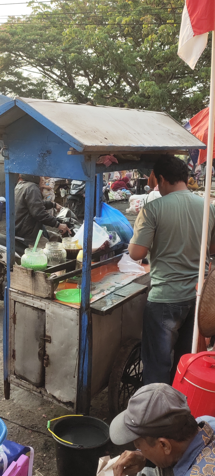 In Karangtowo, Indonesia, a street vendor is preparing drinks from a mobile cart. The cart is constructed of wood and metal, with a blue-painted wooden frame and a metal body. On top of the cart, there are glass jars filled with liquids, one green and two brown. A person in a green t-shirt and blue jeans stands behind the cart, facing away from the camera, and appears to be serving a customer or preparing ingredients. In the foreground, a man wearing a gray baseball cap and a patterned shirt is seated, looking down at something in his hands, possibly food. To the left of the cart, a person in a dark jacket is seated on a motorcycle. The background reveals trees, utility poles, and other market stalls, indicating a busy outdoor environment. A red cooler and a black bucket filled with water are also visible near the cart. An Indonesian flag is partially visible on the right side of the image. The lighting suggests it is late afternoon or early evening.