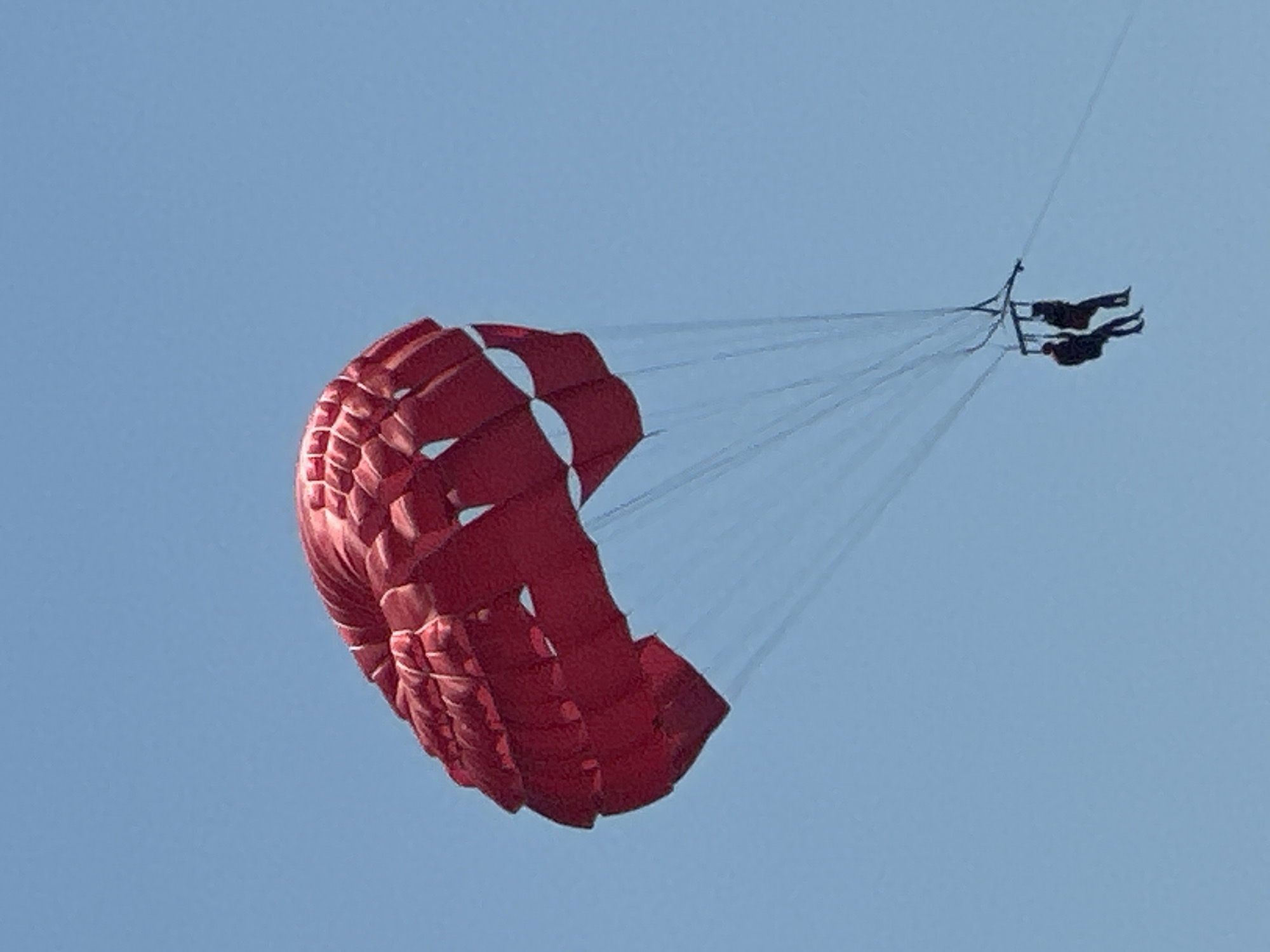 The media file shows two people parasailing against a clear blue sky. The parasail is predominantly red with white sections, and it is inflated and appears to be catching the wind. The two individuals are strapped into a harness, positioned below the parasail, and are being pulled through the air. Their bodies are horizontal, suggesting they are enjoying the ride.
The scene is set during the daytime, as indicated by the bright, cloudless sky. There are no obvious location cues other than the sky itself. The overall impression is one of a recreational activity, likely a tourist attraction, taking place in an open-air environment, potentially over water given the nature of parasailing. The lighting suggests sunny weather. No text is visible in the image. The activity is parasailing, which involves being towed by a boat while attached to a parachute-like canopy. The individuals appear to be experiencing a thrill or enjoying the aerial view.