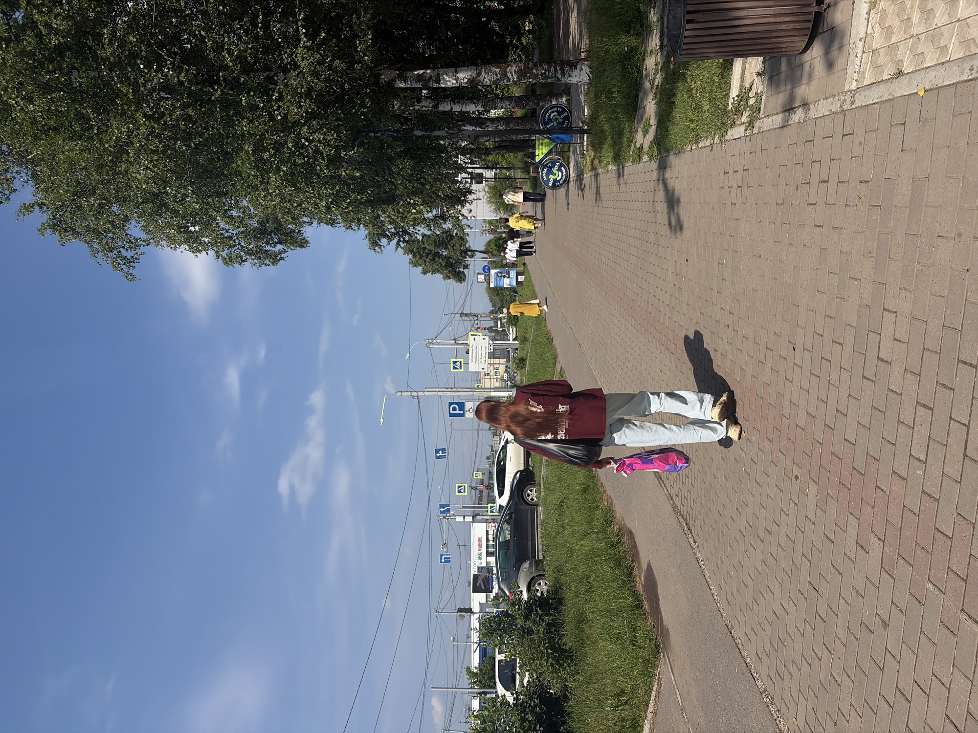 A young woman with long auburn hair walks away from the camera down a brick-paved sidewalk in Chaikovsky, Russia. She wears a dark maroon sweatshirt with white lettering and light blue pants. She carries a large black tote bag over her left shoulder and a bright pink and purple shopping bag in her right hand. The sidewalk is bordered by a grassy area on the left and trees on the right. In the background, several cars are parked and driving on a road. Traffic signals and streetlights are visible, along with buildings and overhead power lines. Other pedestrians are walking further down the sidewalk, some wearing bright clothing. The sky is blue with scattered white clouds.