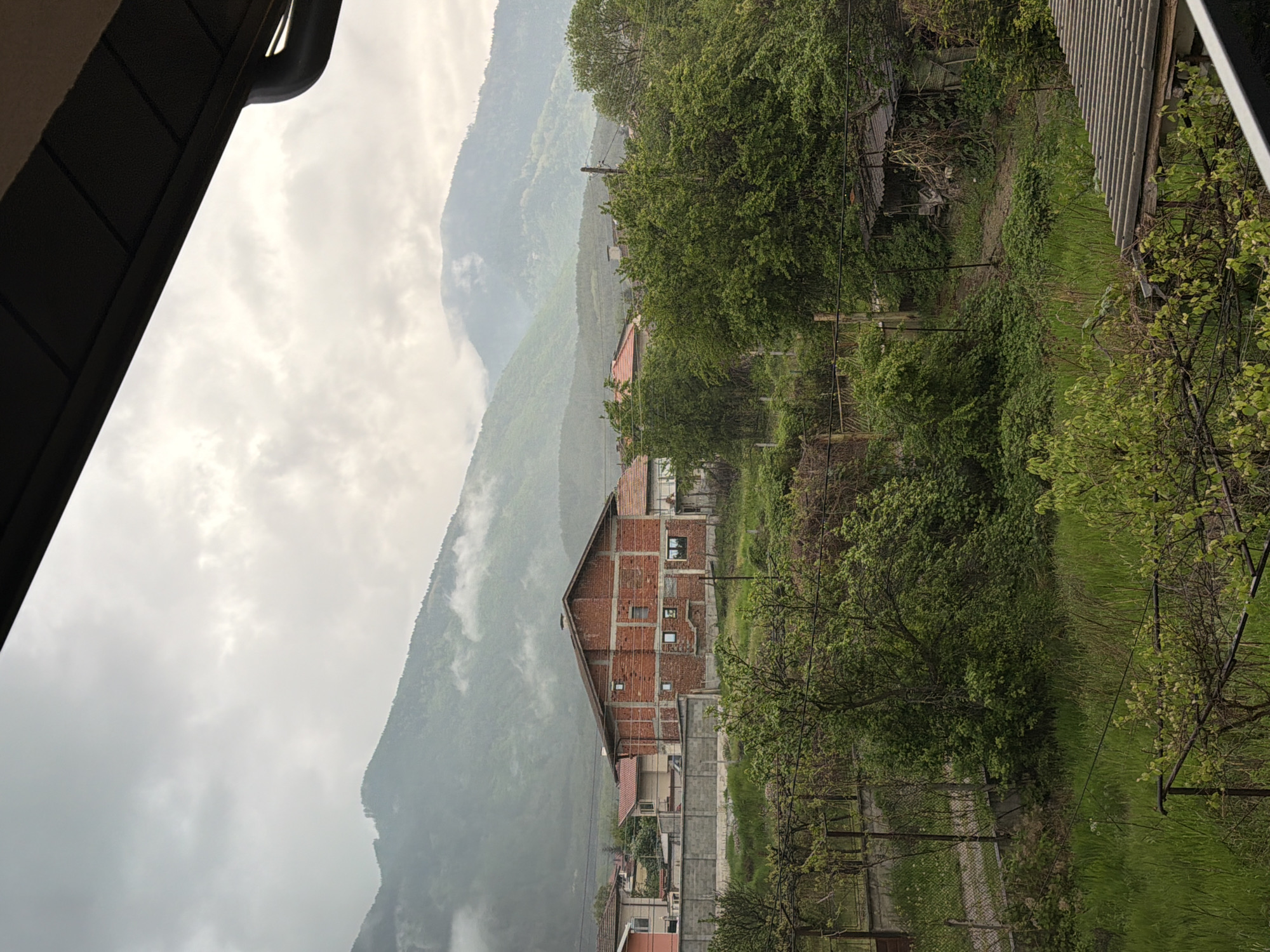 The image depicts a mountainous landscape near Resilovo, Bulgaria, under overcast skies. A partially constructed brick building is visible in the foreground, with a mountain range rising in the background, partly obscured by mist. Vegetation is prominent, with trees and grass covering the hillside. Adjacent to the unfinished brick structure, other residential buildings are visible. Power lines stretch across the frame, and elements of architecture are visible at the top and right of the frame.