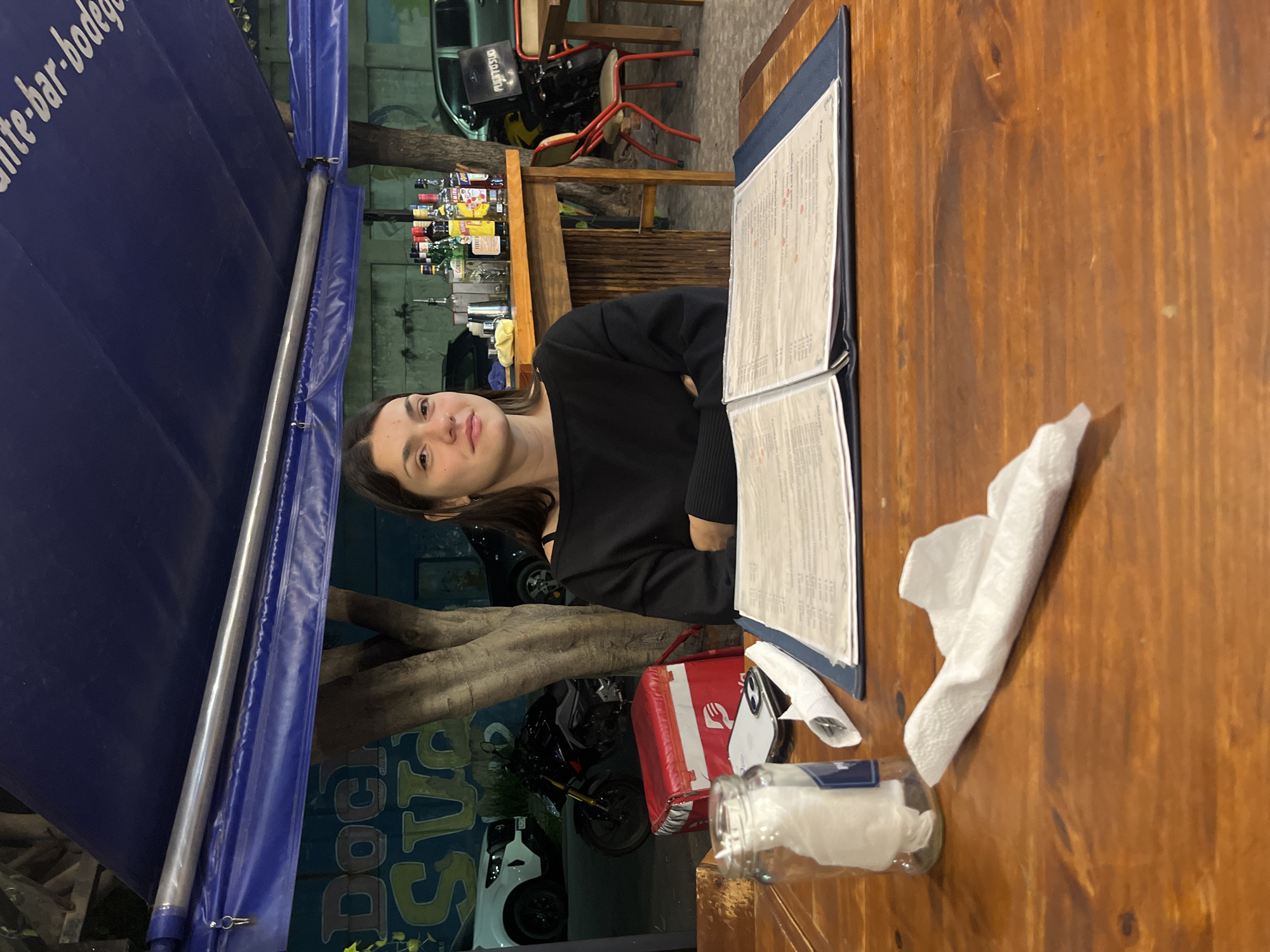 The image depicts a young woman sitting at a wooden table in an outdoor dining area. She is positioned next to a menu and a container with napkins. A red delivery box is also on the table. The restaurant appears to be in Dock Sud, Argentina, indicated by a blue awning with the word "Dock" visible. The background features a bar with various bottles and a delivery motorcycle.