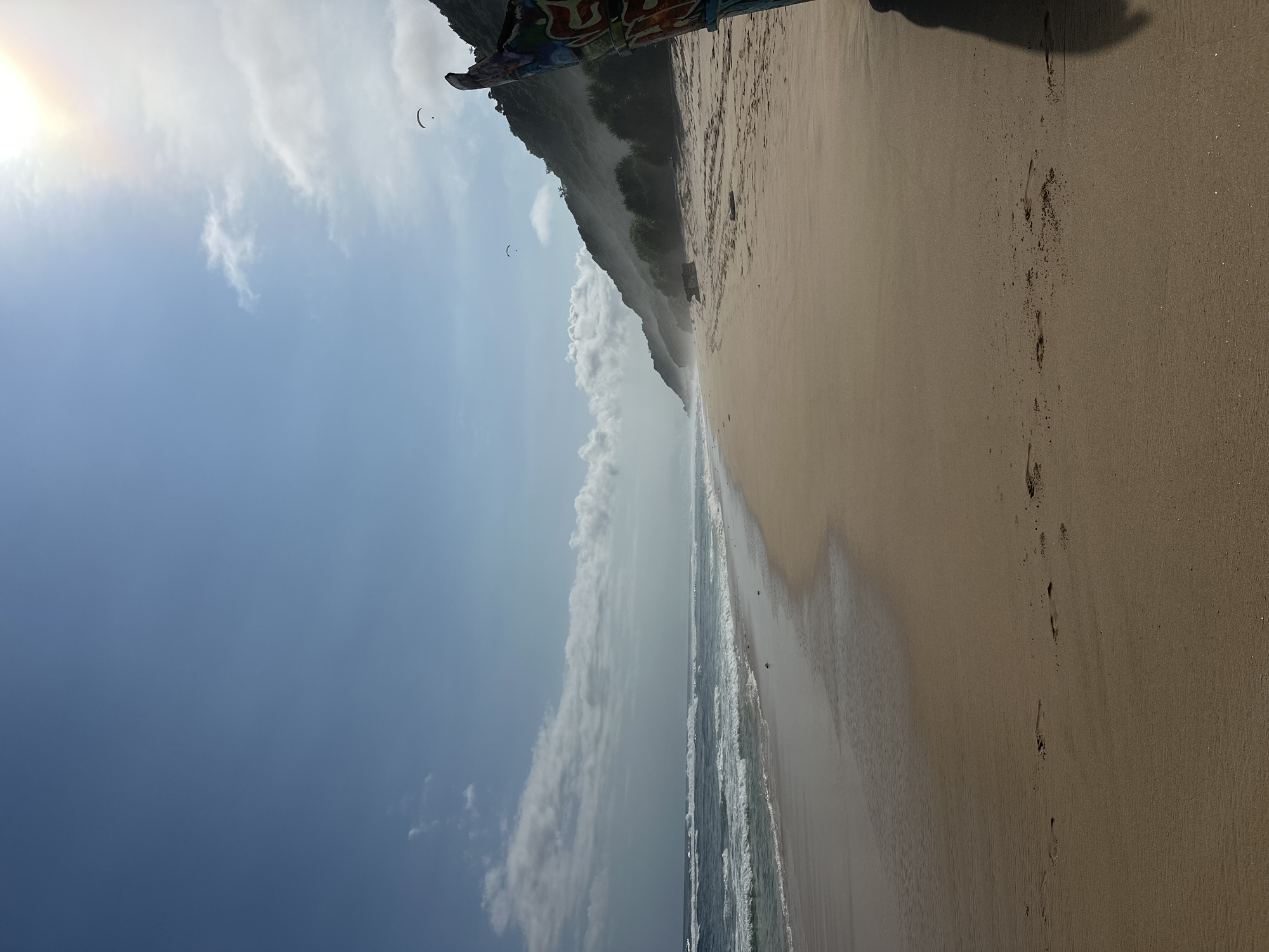 The image shows a beach scene in Kuta Selatan, Indonesia, featuring a sandy beach meeting the ocean on the right side of the image. The sky occupies the left portion of the image, displaying blue skies with clouds and the sun. A headland or cliff covered in vegetation is seen behind the beach. Two paragliders or similar aerial crafts are visible in the sky. There are footprints in the sand. A shadow is cast in the upper right corner of the image, likely from the photographer.