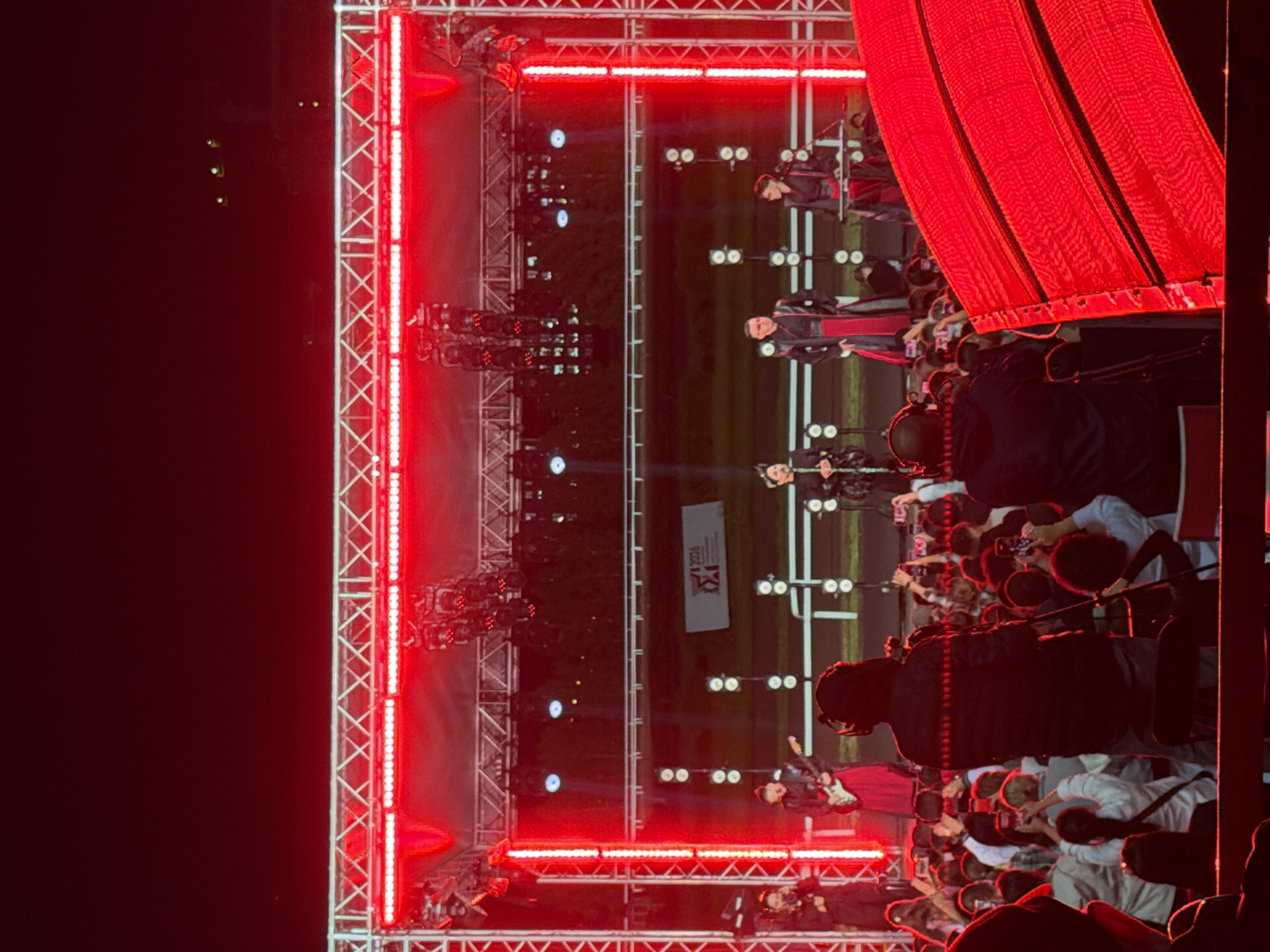 An outdoor night concert is in progress, with a stage set up and bathed in red lighting. A line of performers in traditional attire stands on the stage. The audience is visible in the foreground, many holding up phones to capture the event. A large screen, also illuminated in red, is positioned in the lower right portion of the frame. The scene suggests a lively and engaging performance, with a clear focus on the spectacle of the stage and the enthusiasm of the crowd.